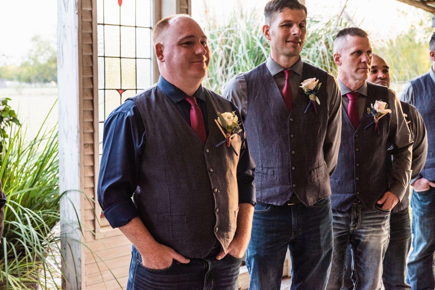 Three groomsmen in navy vests and red ties stand together at a wedding venue.