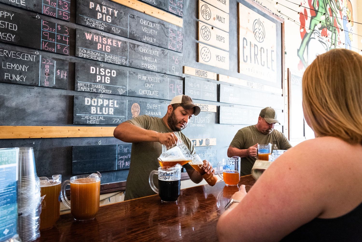A bartender serves drinks at a craft brewery with chalkboard menus on the wall.