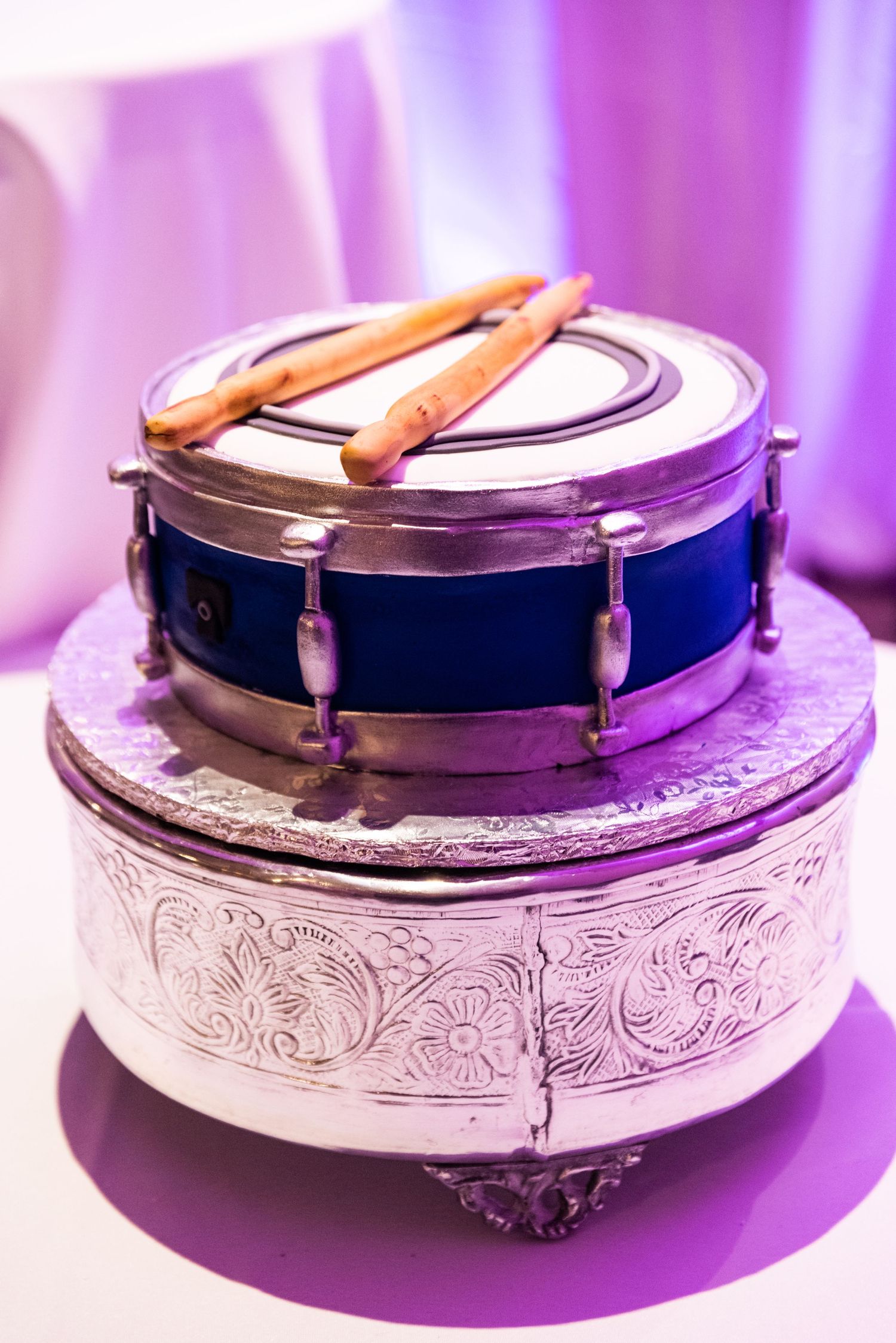 A snare drum placed on top of an ornate silver cake stand under purple lighting.