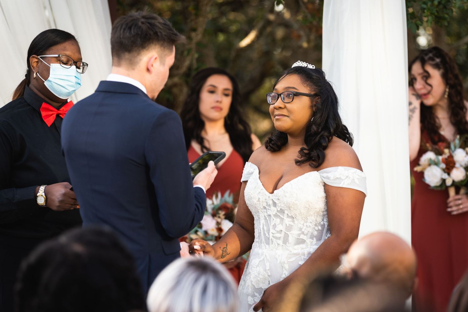 A wedding ceremony takes place under a white draped arch with the officiant in a red bowtie and black mask.