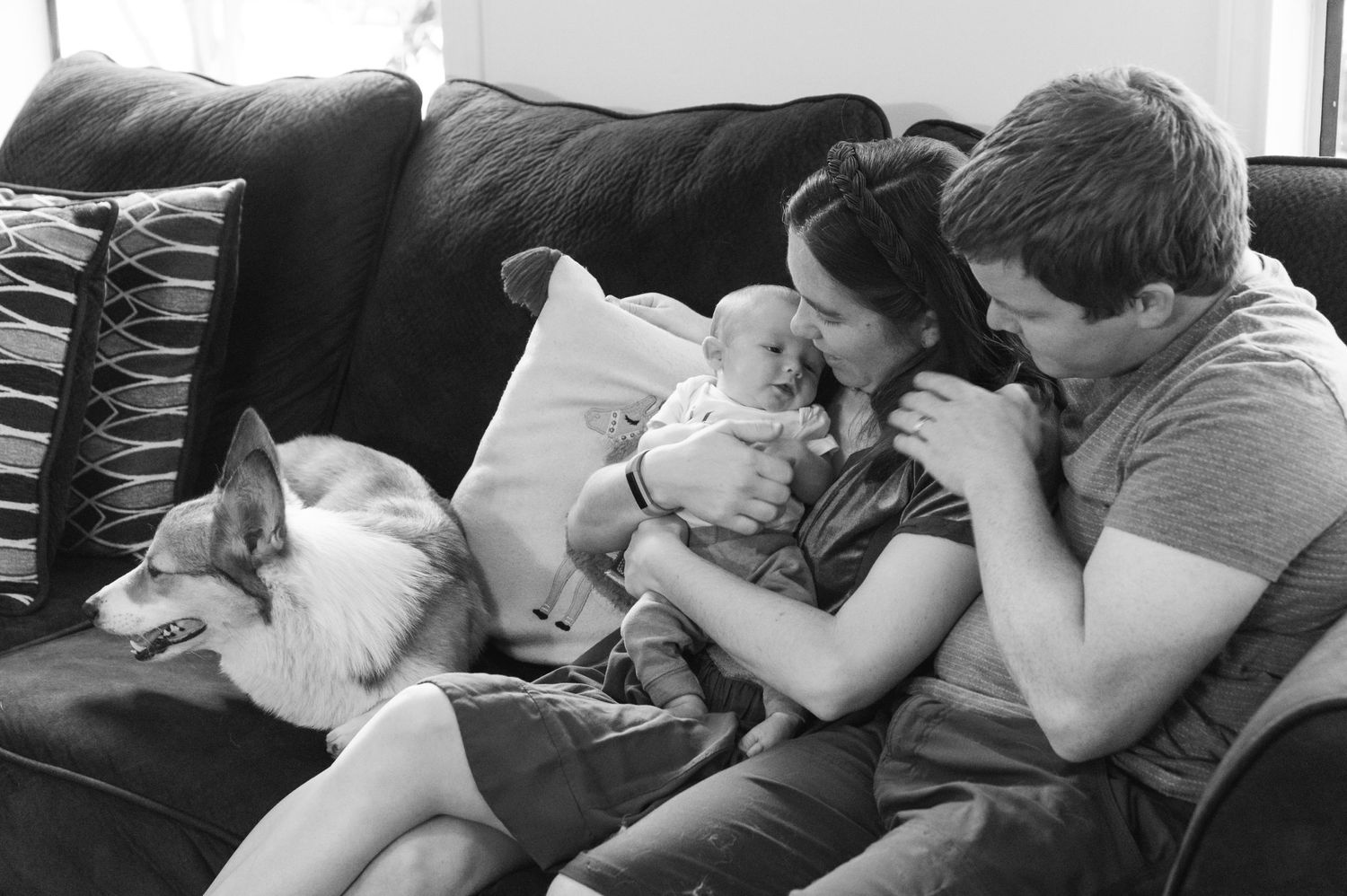 Couple cuddles with baby on couch while cat sleeps nearby in black and white photo.
