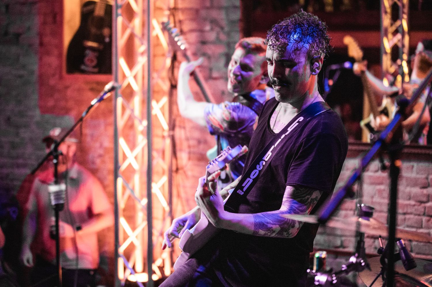 Musicians perform on stage with atmospheric blue lighting against exposed brick wall.