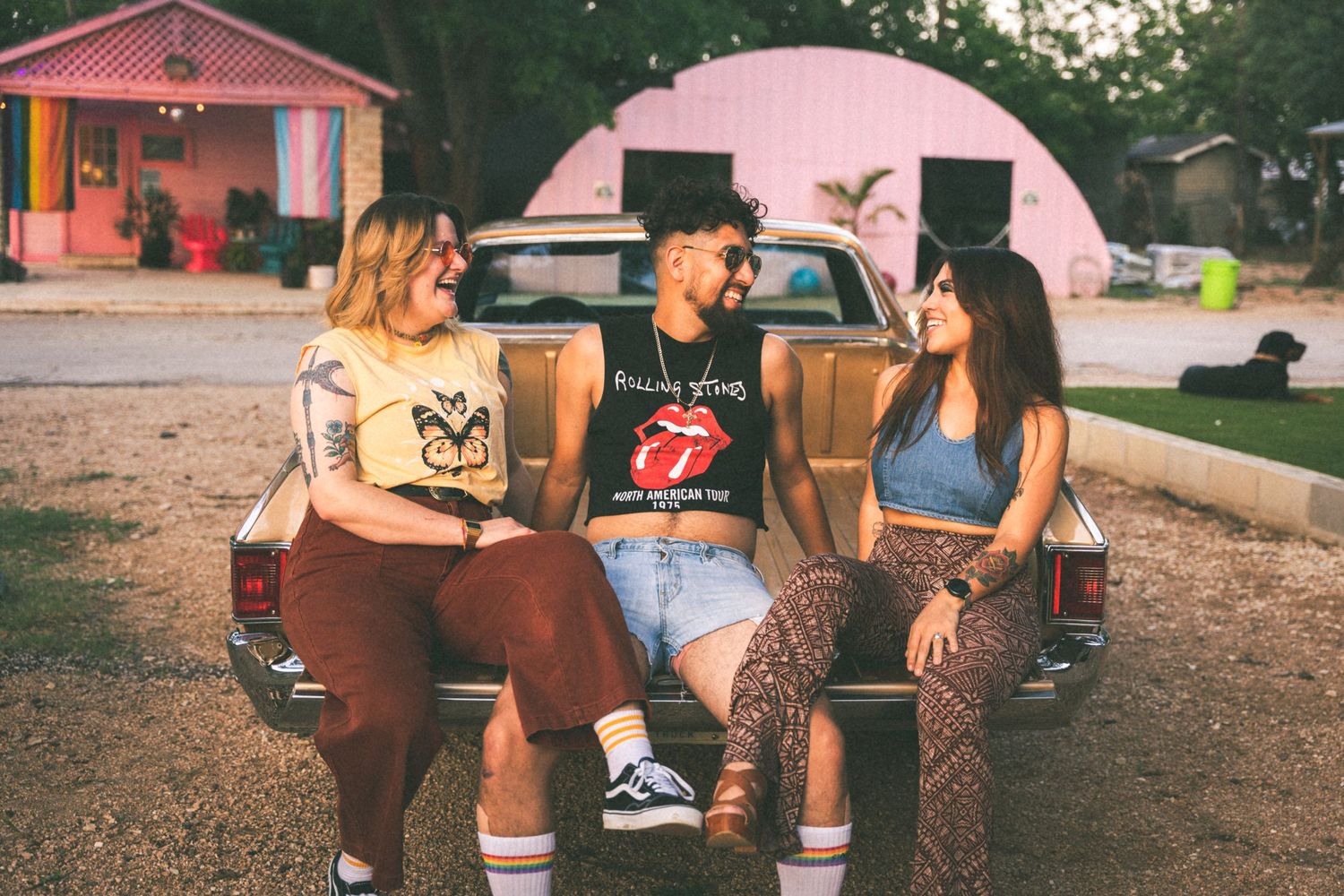 Three friends sit on truck tailgate having conversation at sunset near pink building.