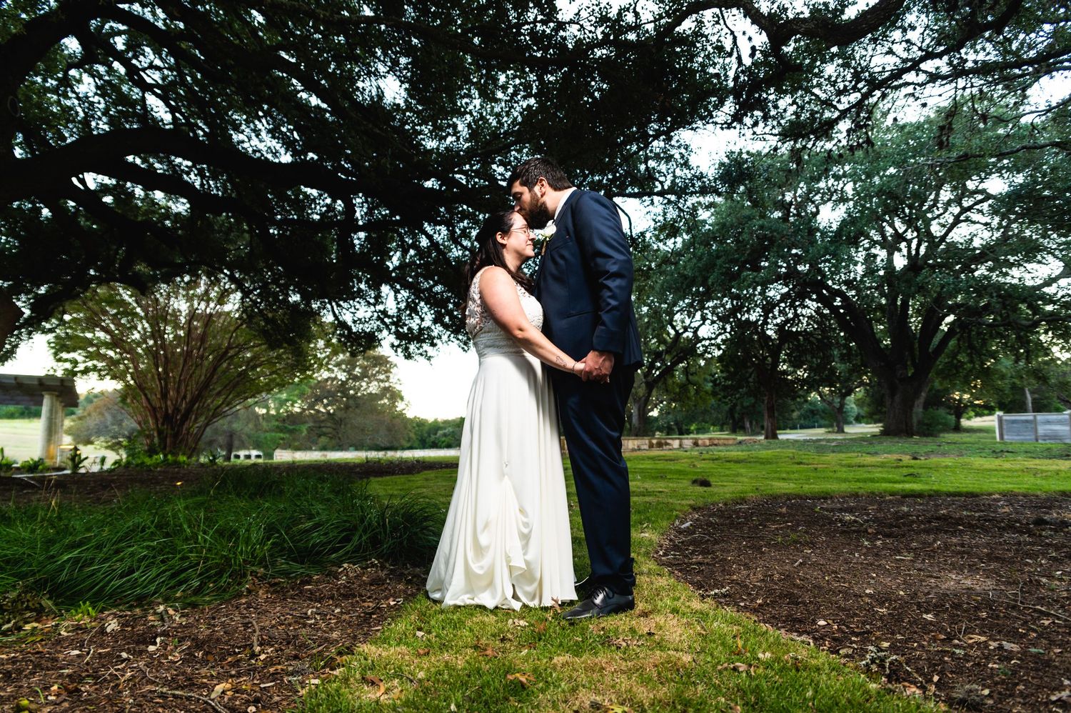 A romantic moment between newlyweds standing under evergreen trees on a grassy lawn.