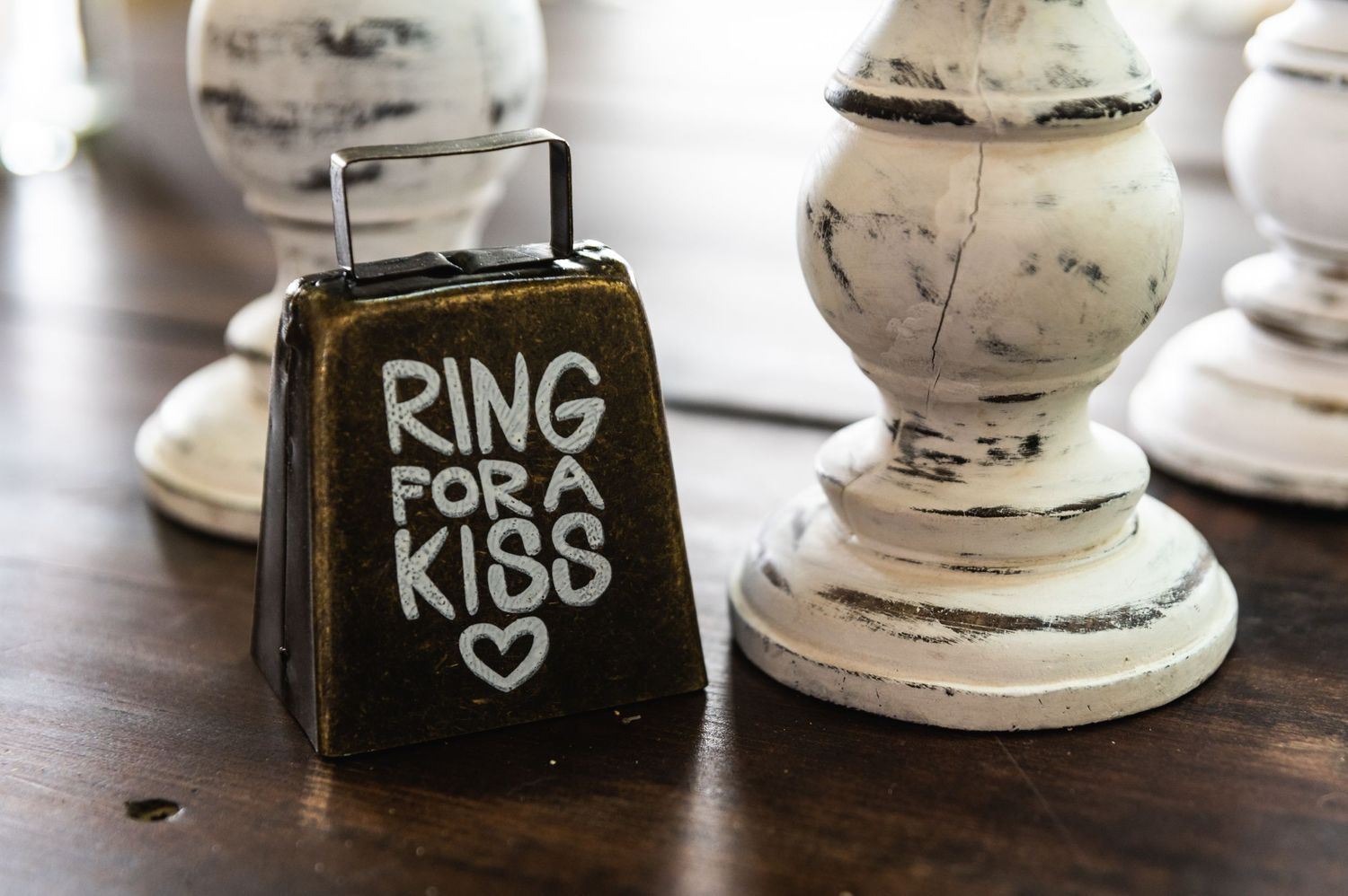 Vintage-style bell with 'Ring for a Kiss' text next to white distressed decorative pillar.