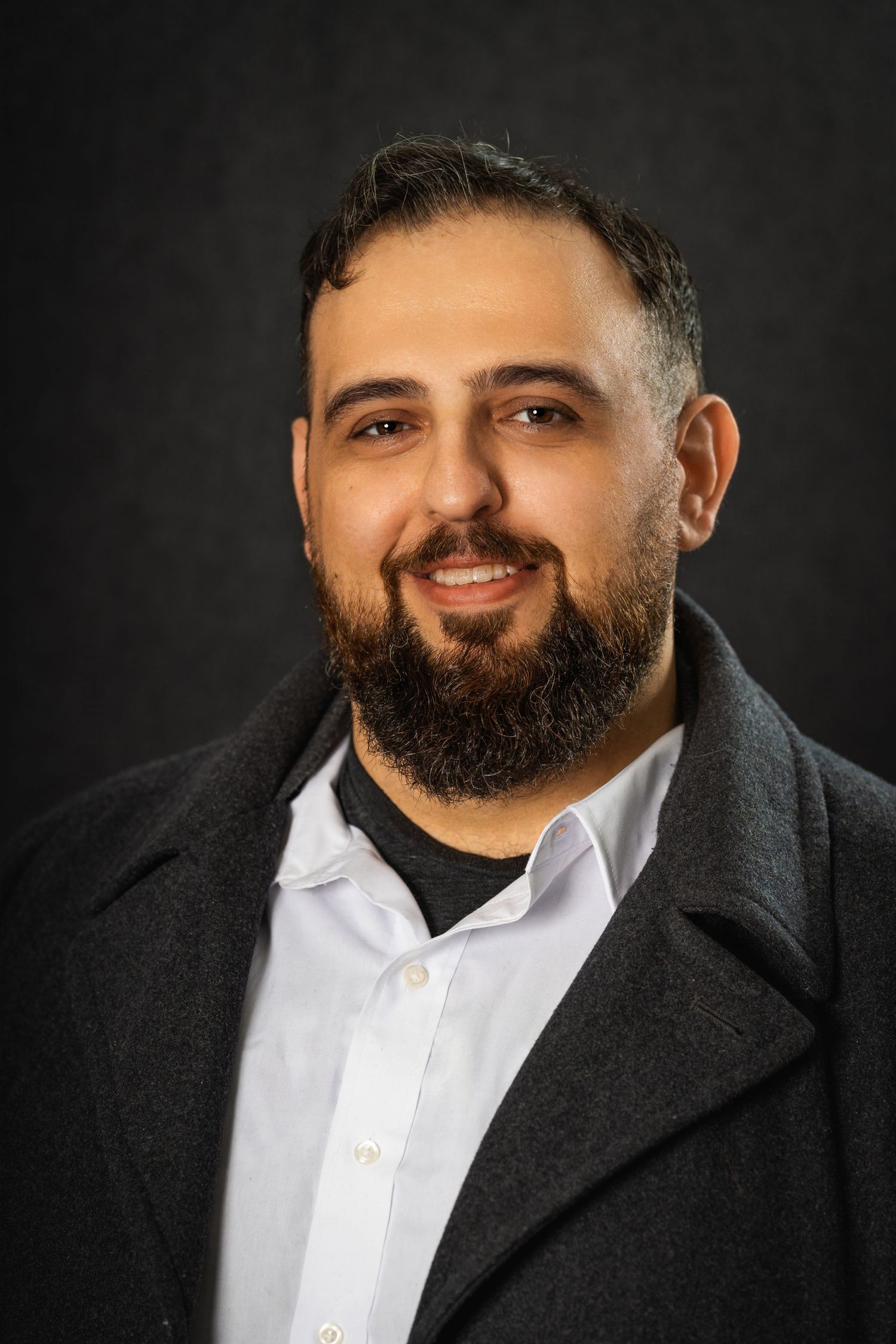 Professional headshot of a bearded man in dark coat and white shirt against black background.