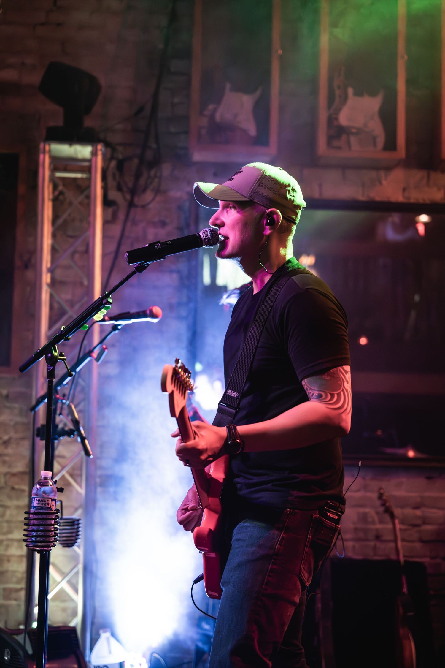 Musician performs with guitar and microphone on stage with colored lighting and brick wall backdrop.