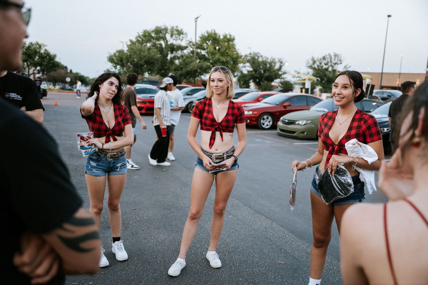 People gathered in a parking lot at a car meet wearing casual summer attire.