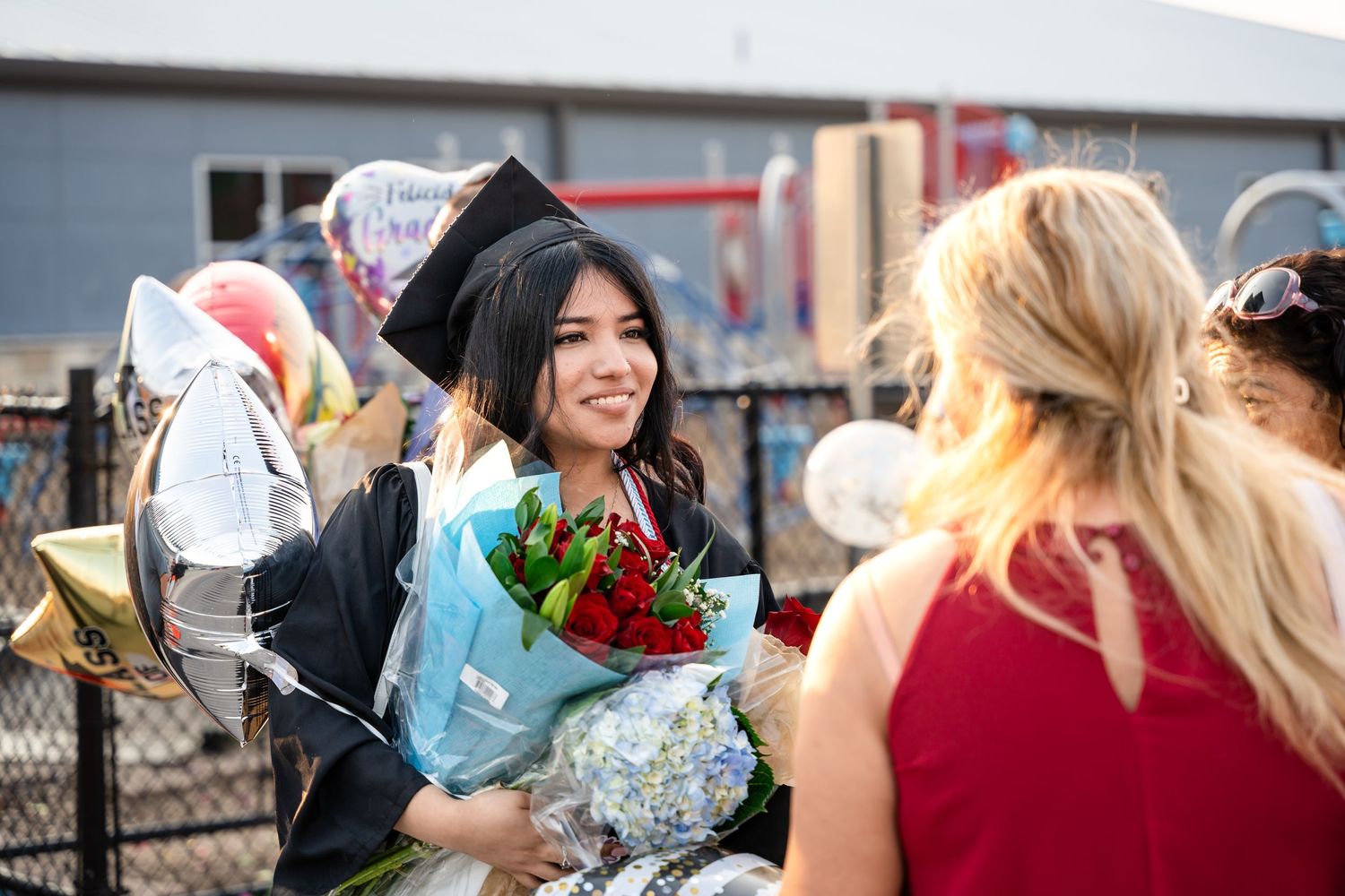 Graduate holding bouquet smiles while celebrating with others outdoors.
