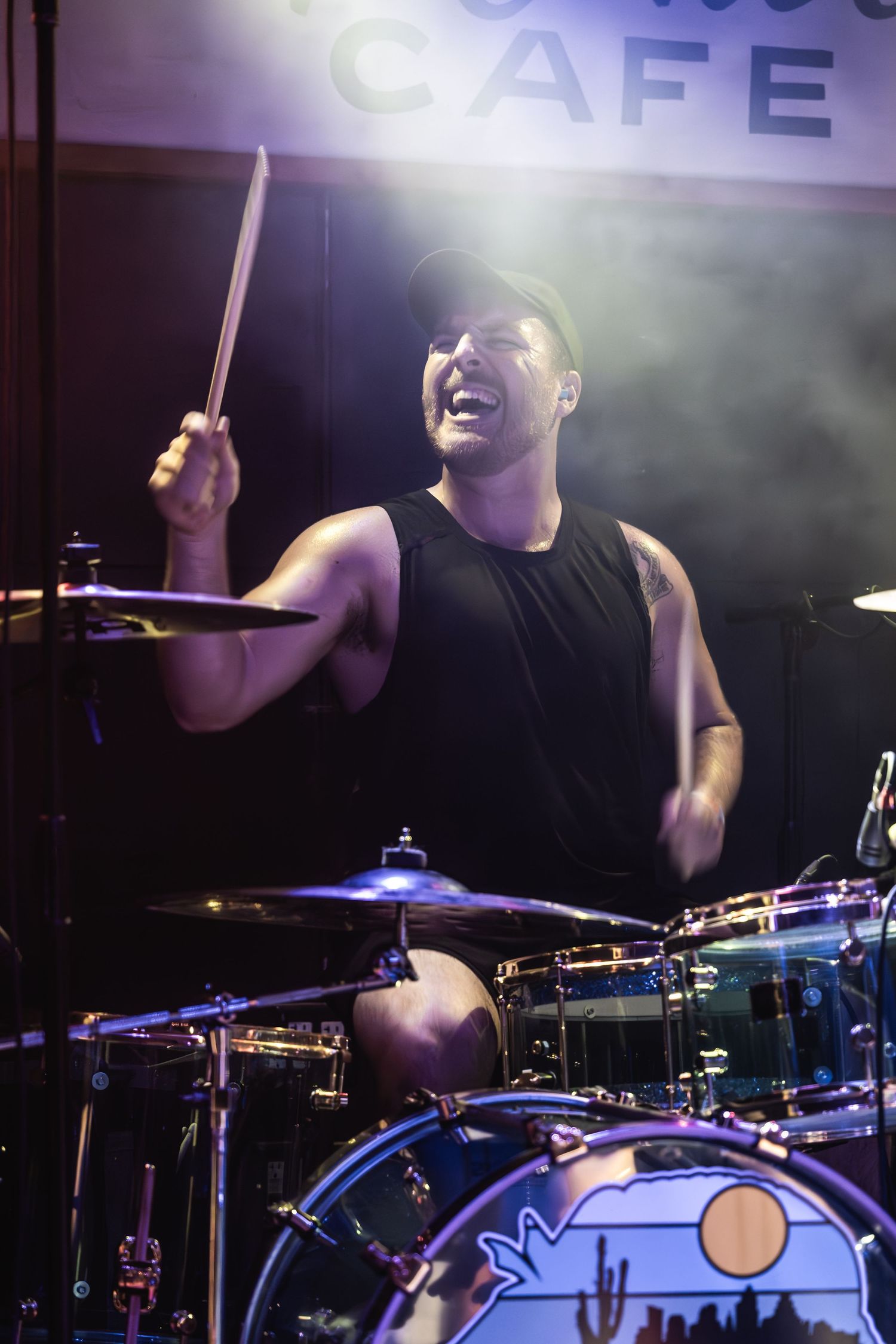 Drummer enthusiastically performs behind kit with illuminated cafe sign visible.