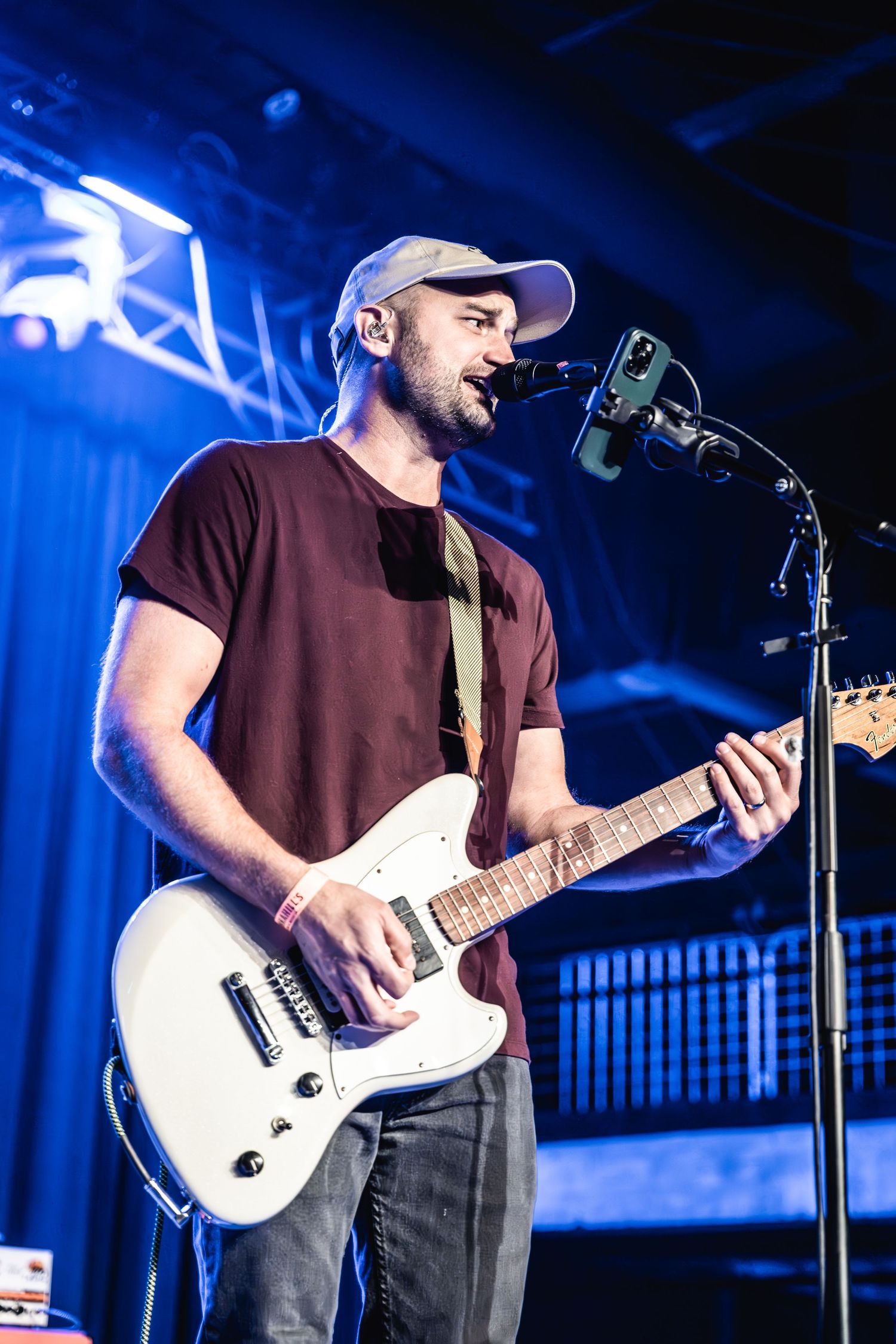 Musician in baseball cap playing white guitar and singing into microphone on stage.