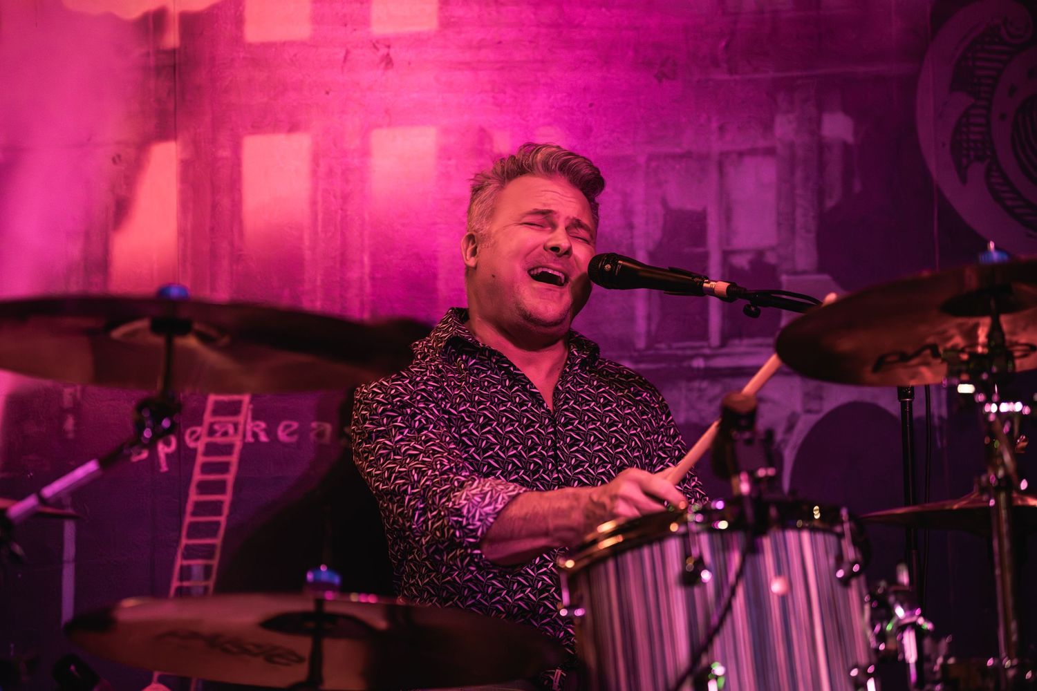 Drummer performs energetically under vibrant pink stage lighting during live performance at Speakeasy venue.