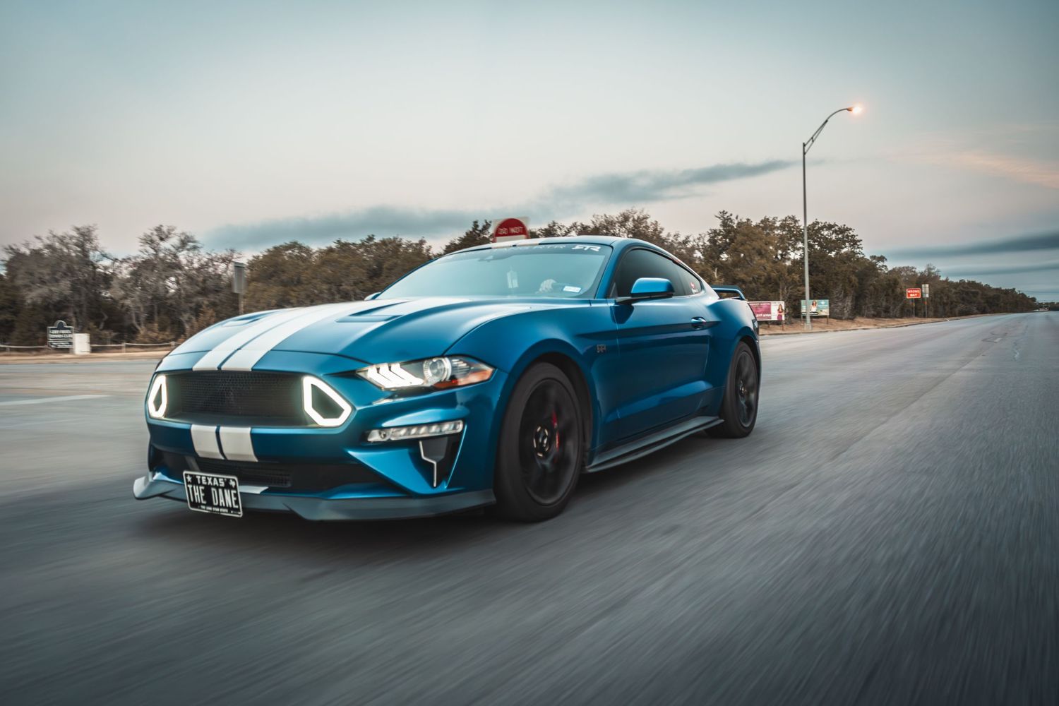 Blue Mustang GT with white racing stripes photographed in motion at dusk.