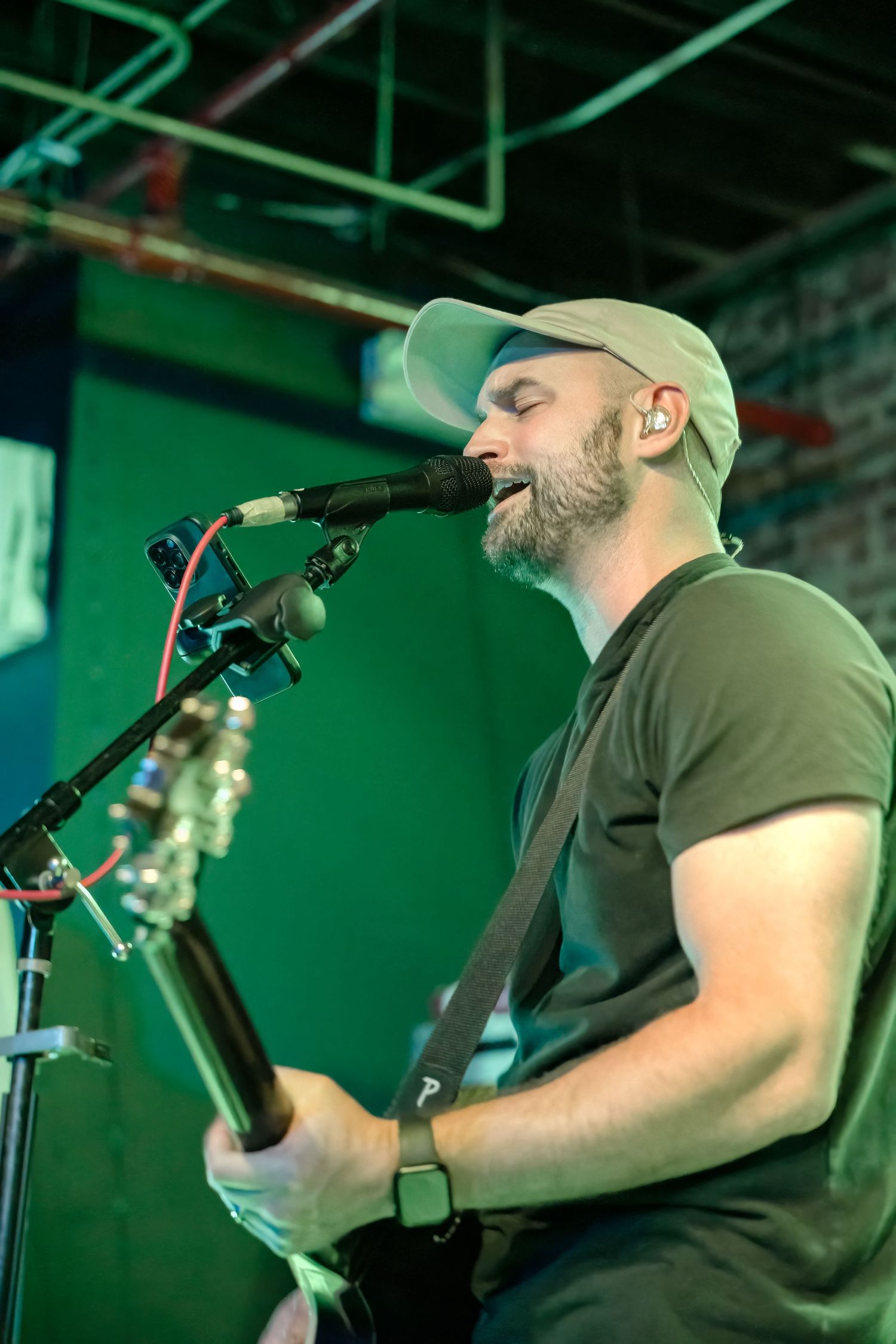 Musician in baseball cap singing into microphone while playing guitar under green lighting.