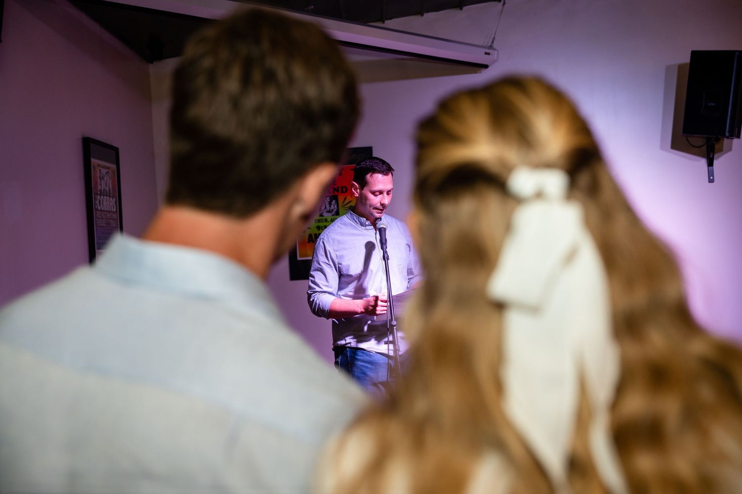 A person speaking at a microphone while others watch in a purple-lit room.