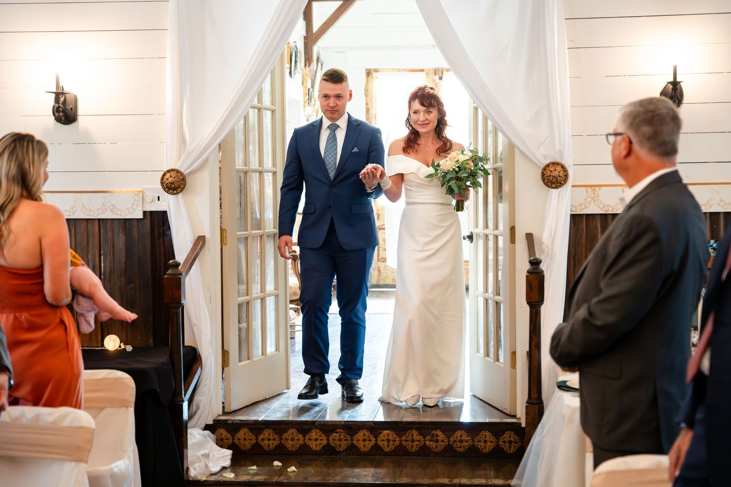 Wedding couple standing at entrance archway decorated with white fabric and wooden accents.