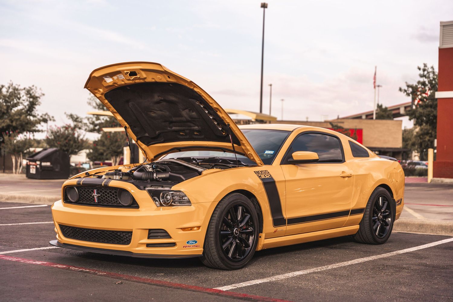 A yellow Ford Mustang Boss 302 with black racing stripes and an open hood sits in a parking lot.