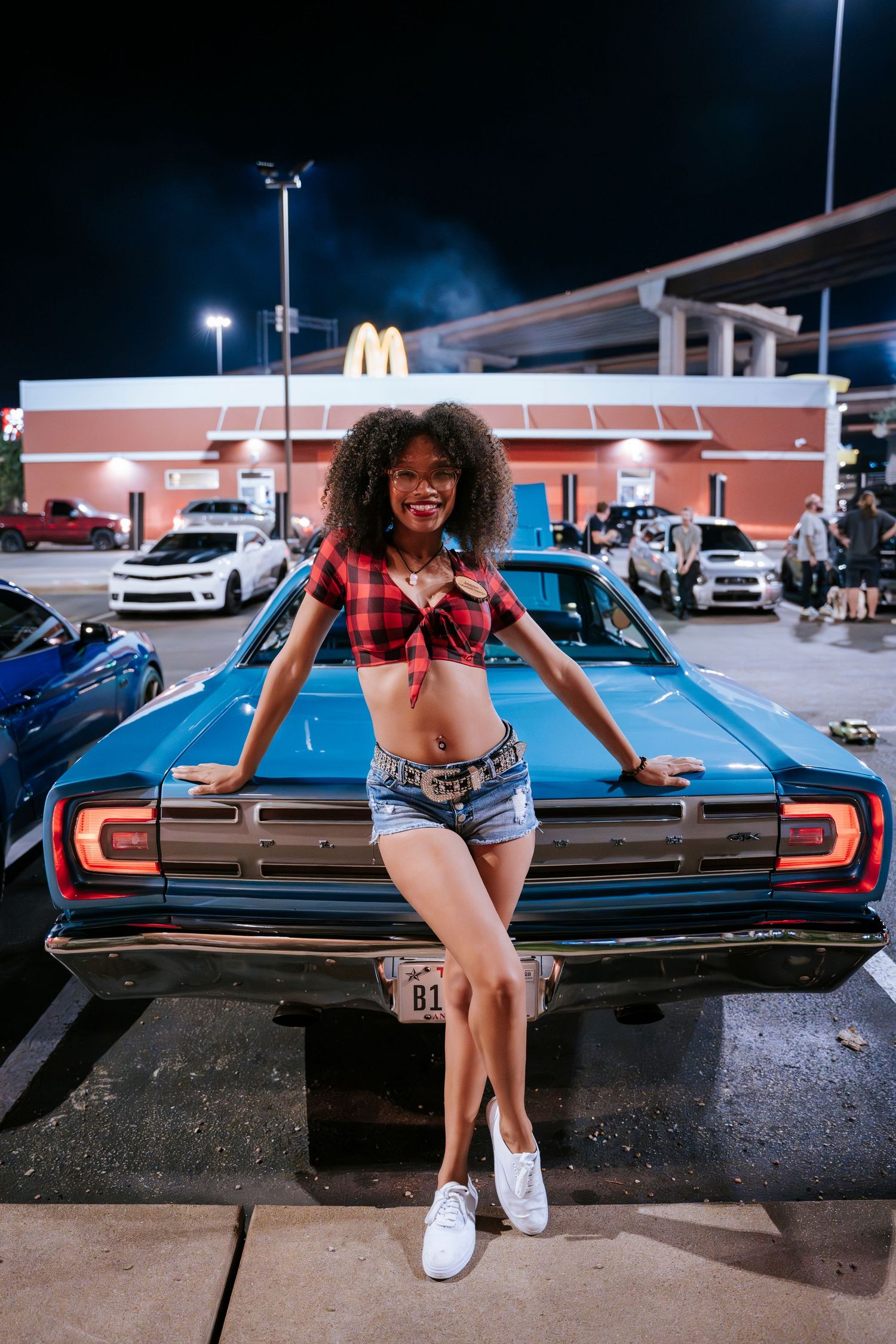 Model in plaid top and denim shorts poses on vintage blue car hood at night.