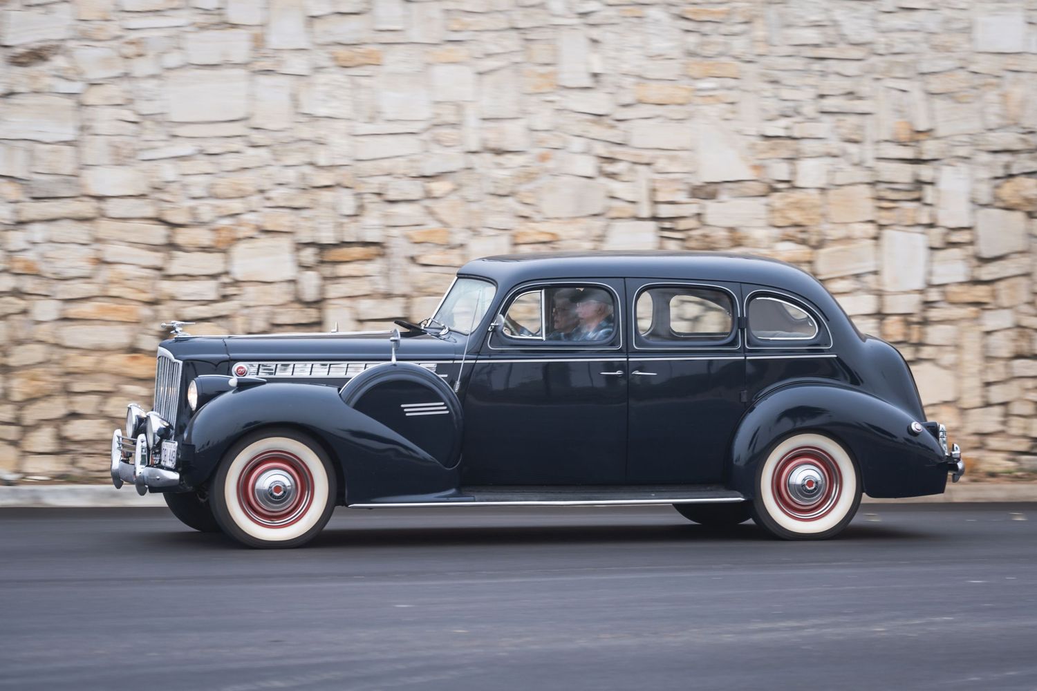 Vintage 1940s Packard sedan in dark blue with white wall tires against stone wall.