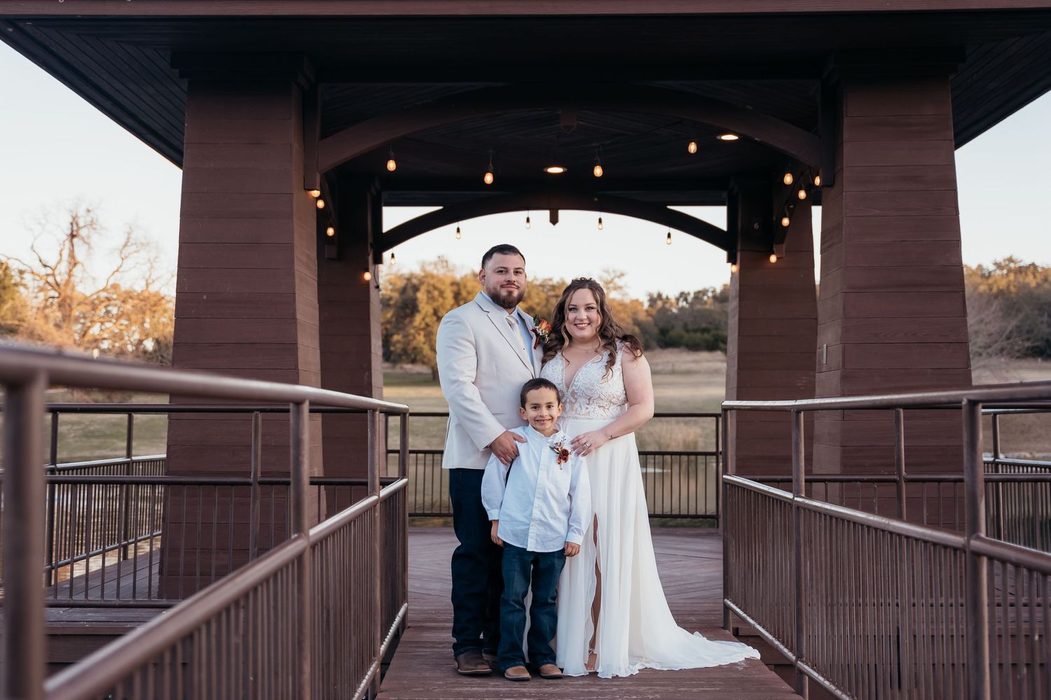 A family of three poses under a covered bridge decorated with string lights.