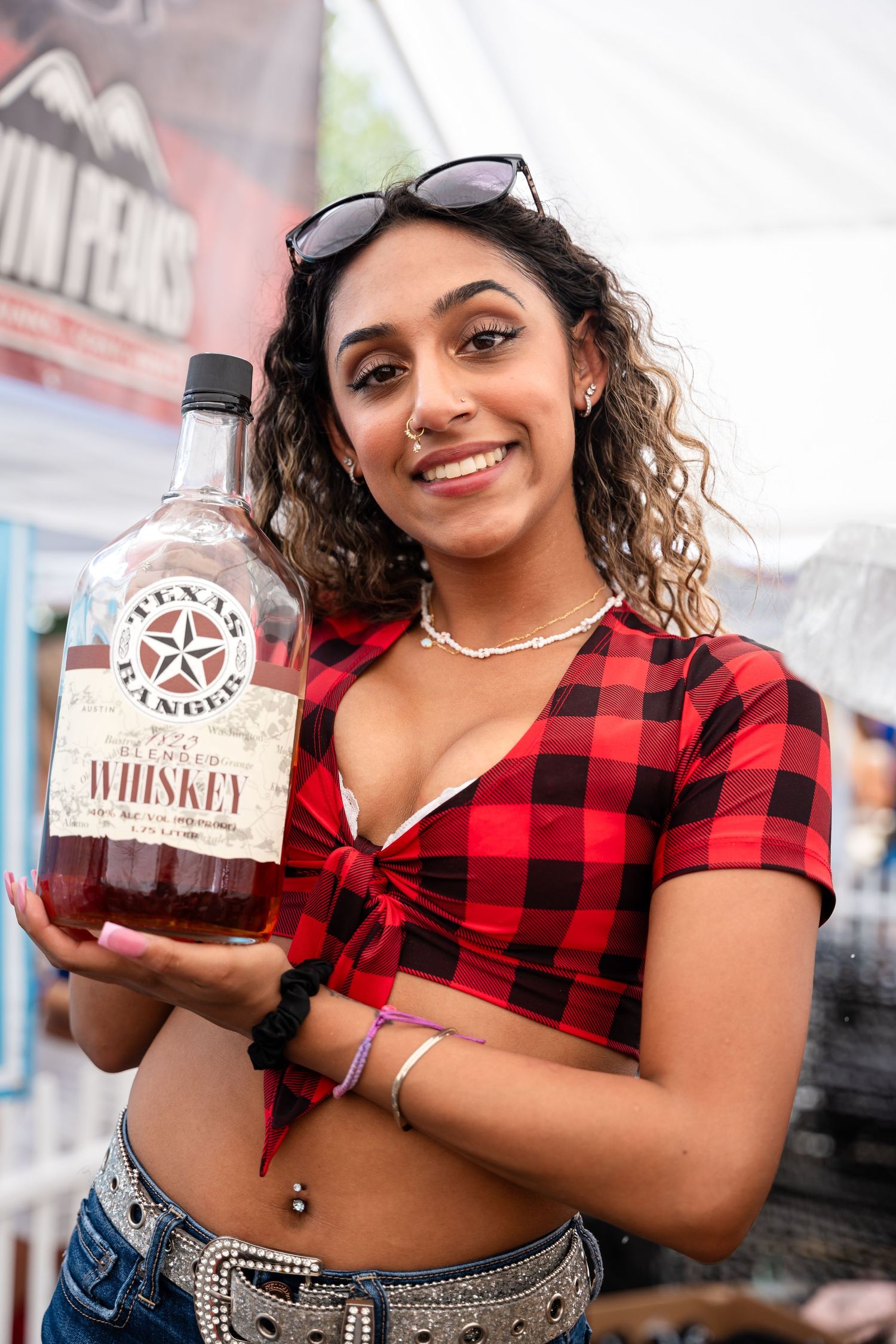Woman in buffalo plaid crop top holding whiskey bottle at festival.