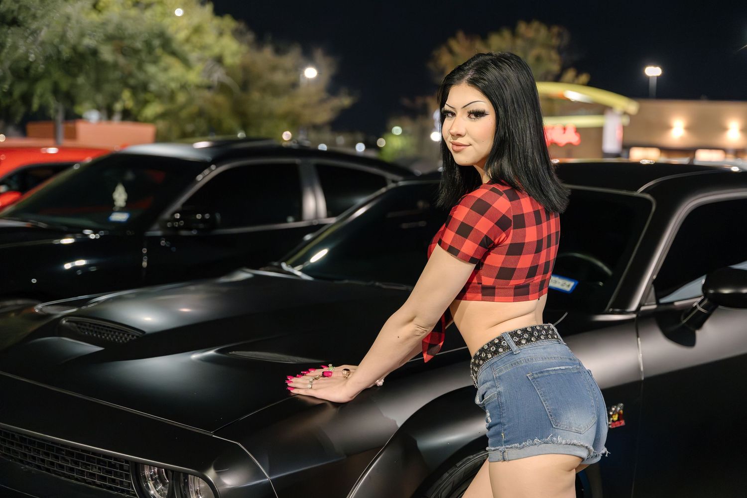 A woman in red plaid crop top and denim shorts poses beside black cars in a nighttime parking lot.