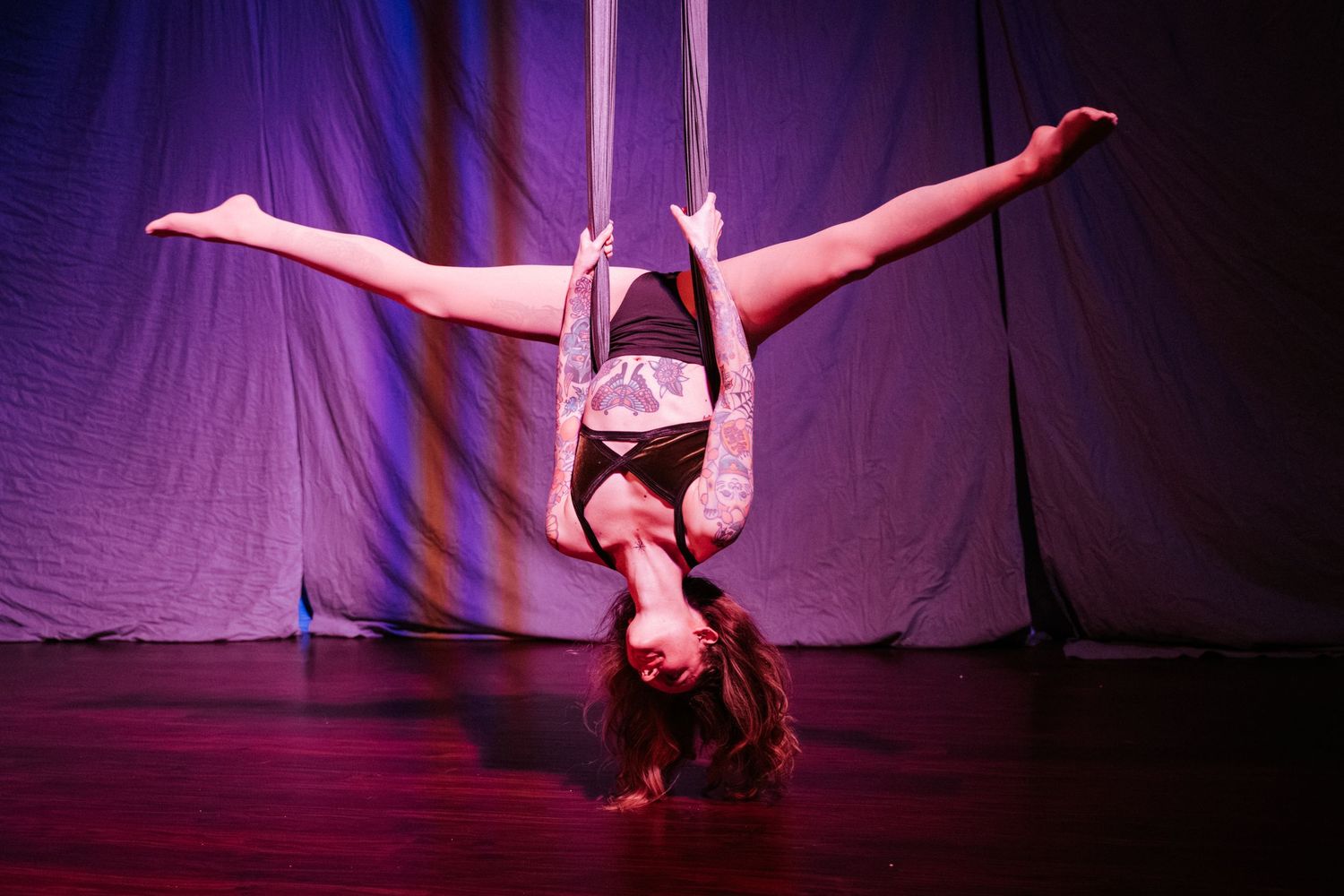 Aerial acrobat performing inverted split pose on silk ribbons on a purple-lit stage.