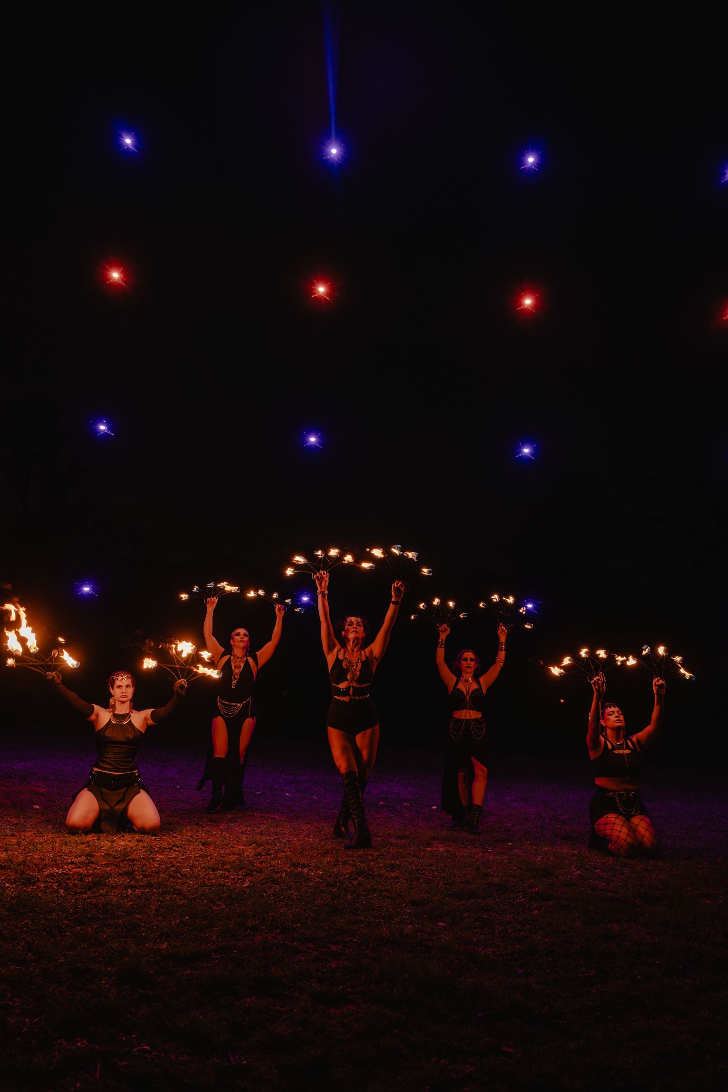 Five female fire performers kneel on grass at night, holding flaming torches aloft under colorful red and blue lights.