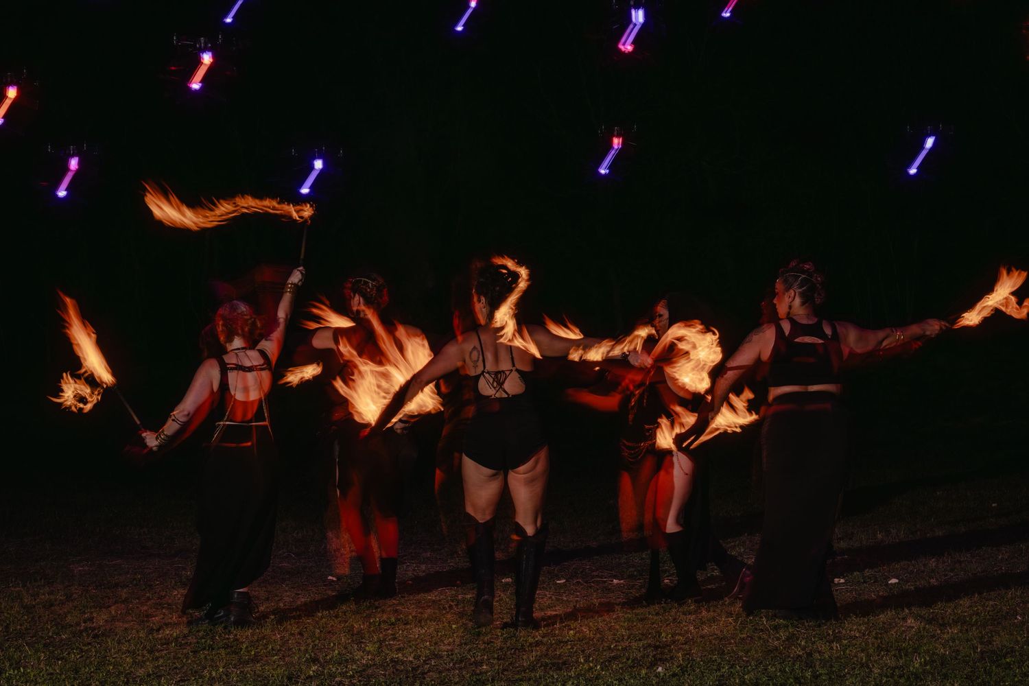 Fire dancers perform at night, wielding flames with glowing light staffs illuminating the dark outdoor stage.