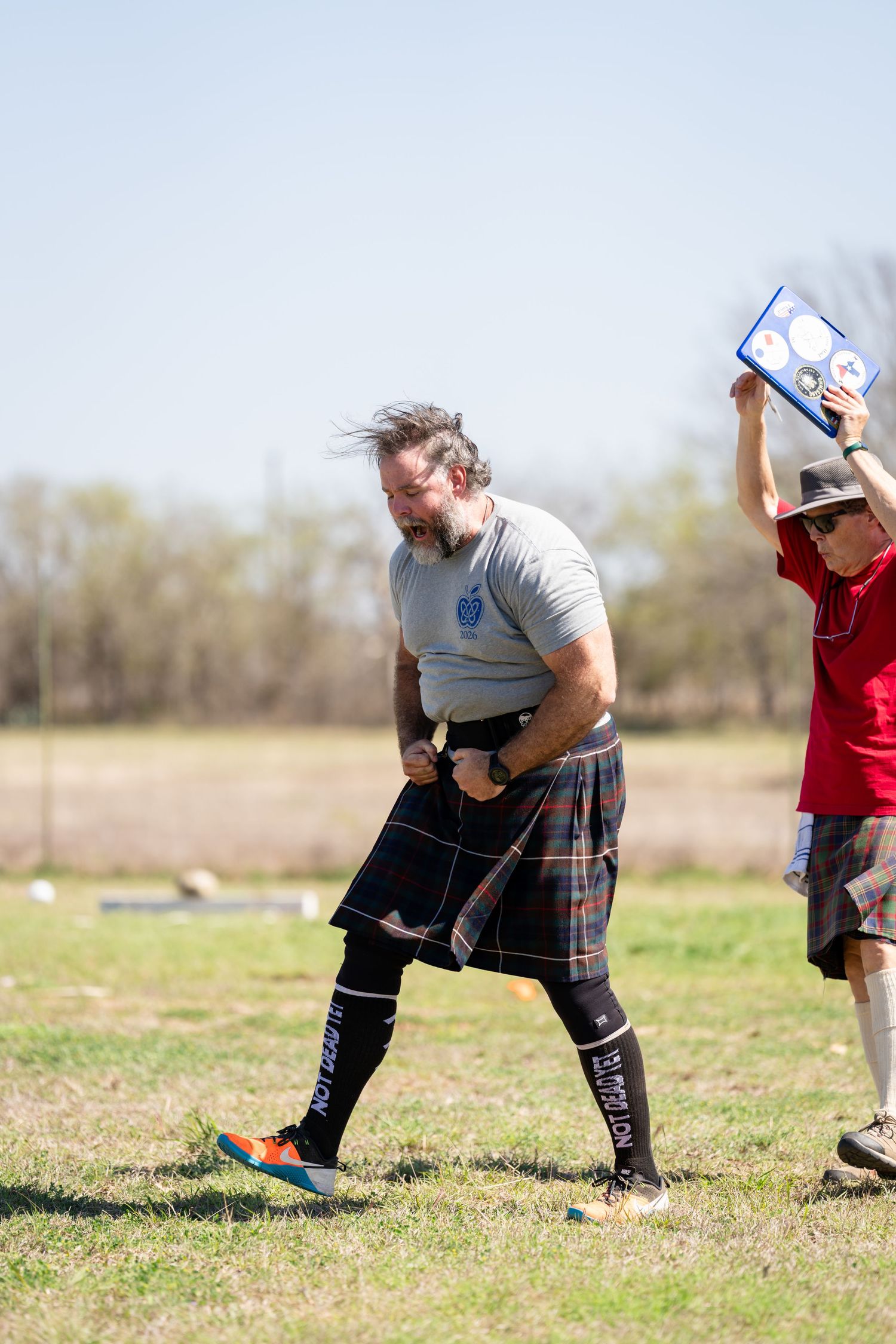 Man in tartan kilt competing at Highland Games event outdoors, with judge holding scoreboard behind him.