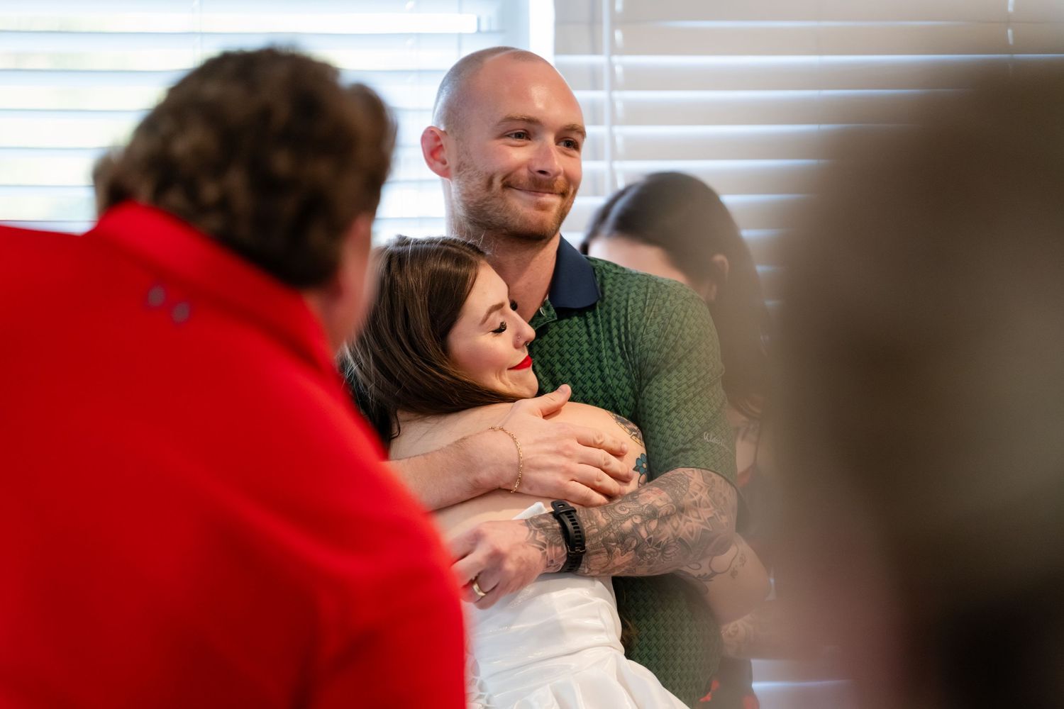 A bald man with tattoos hugs a dark-haired woman in a white dress, smiling warmly indoors.