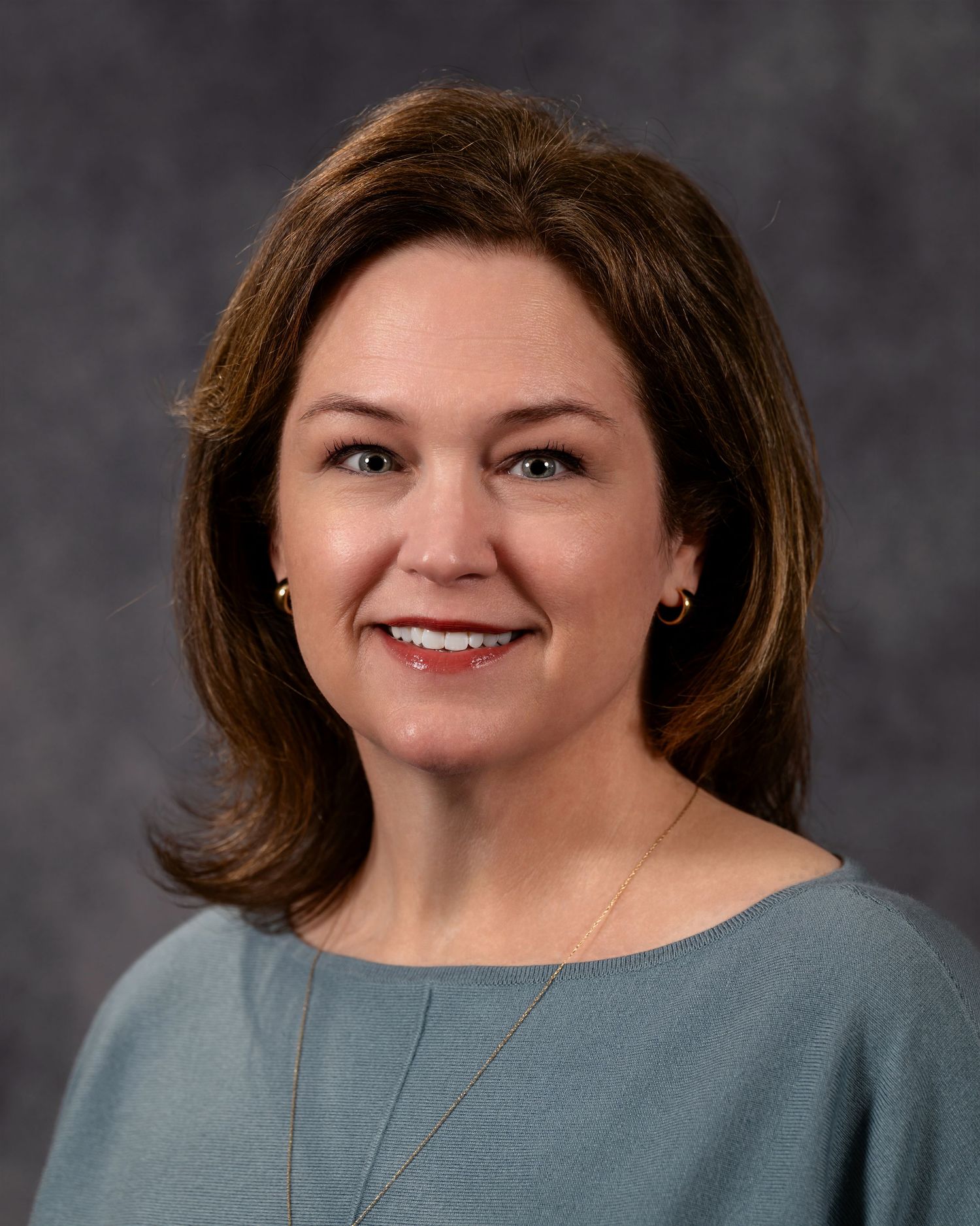 Professional headshot of a smiling woman with brown hair wearing a teal blouse and delicate necklace against a gray background.