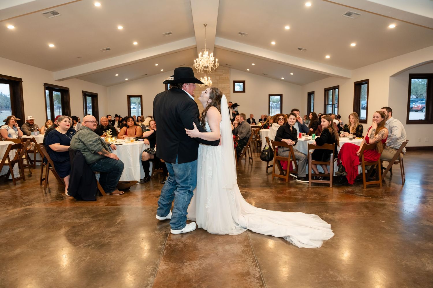 Bride and groom share their first dance at a rustic wedding reception hall filled with seated guests.