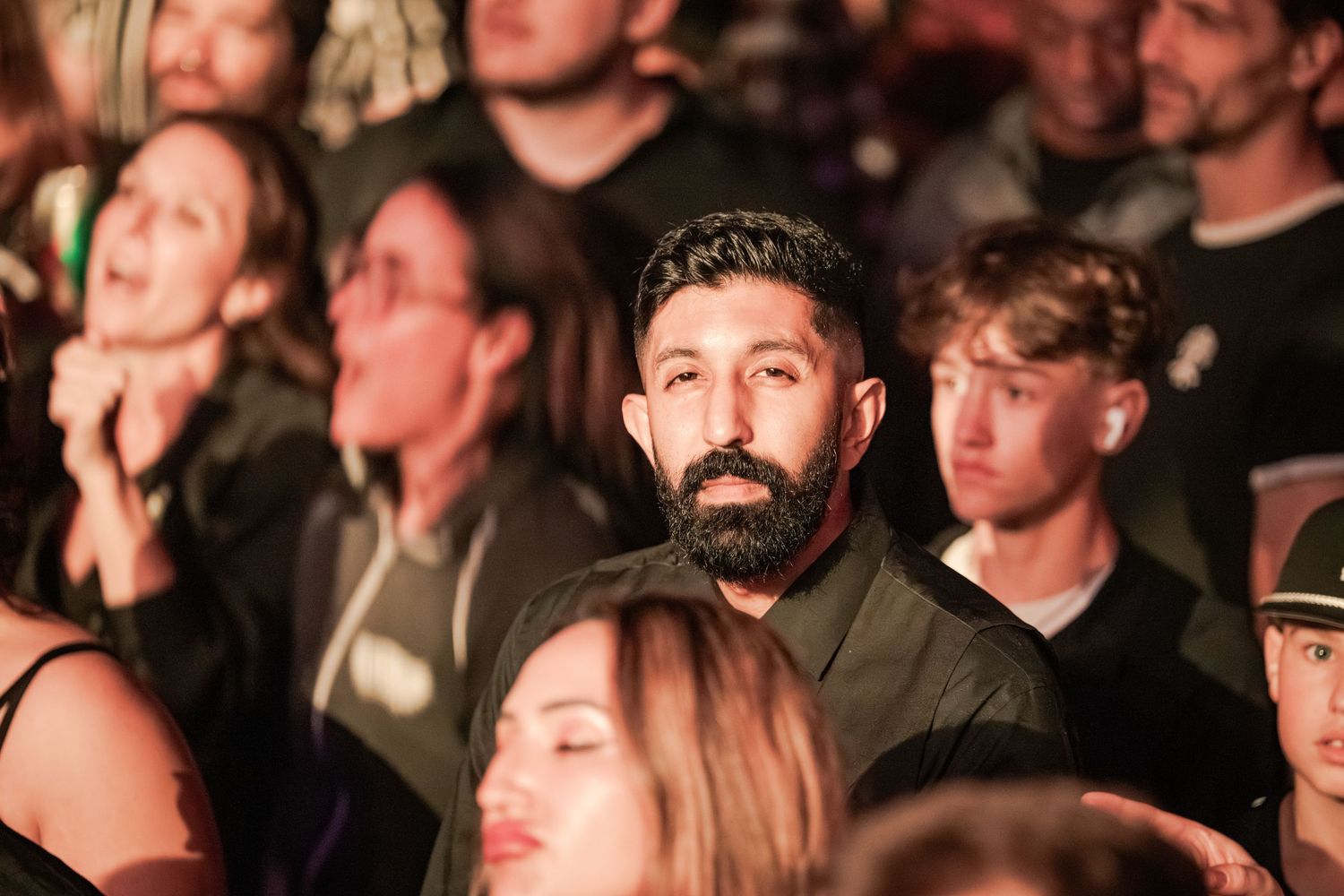 A bearded man in a black shirt stands out in a crowded concert audience, looking directly at the camera.
