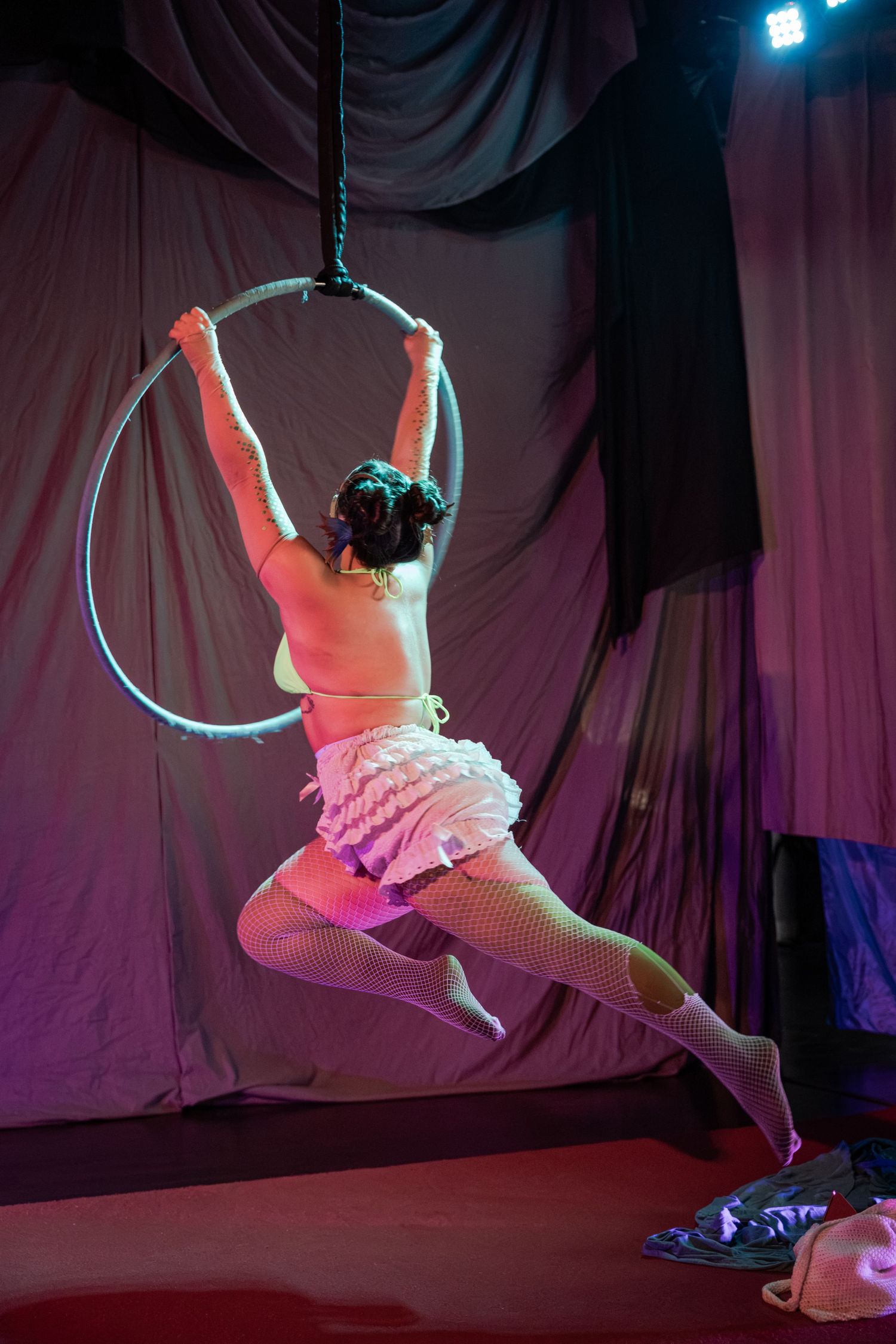 Aerial acrobat performs on a hoop in pink costume and fishnets under stage lighting at a circus.