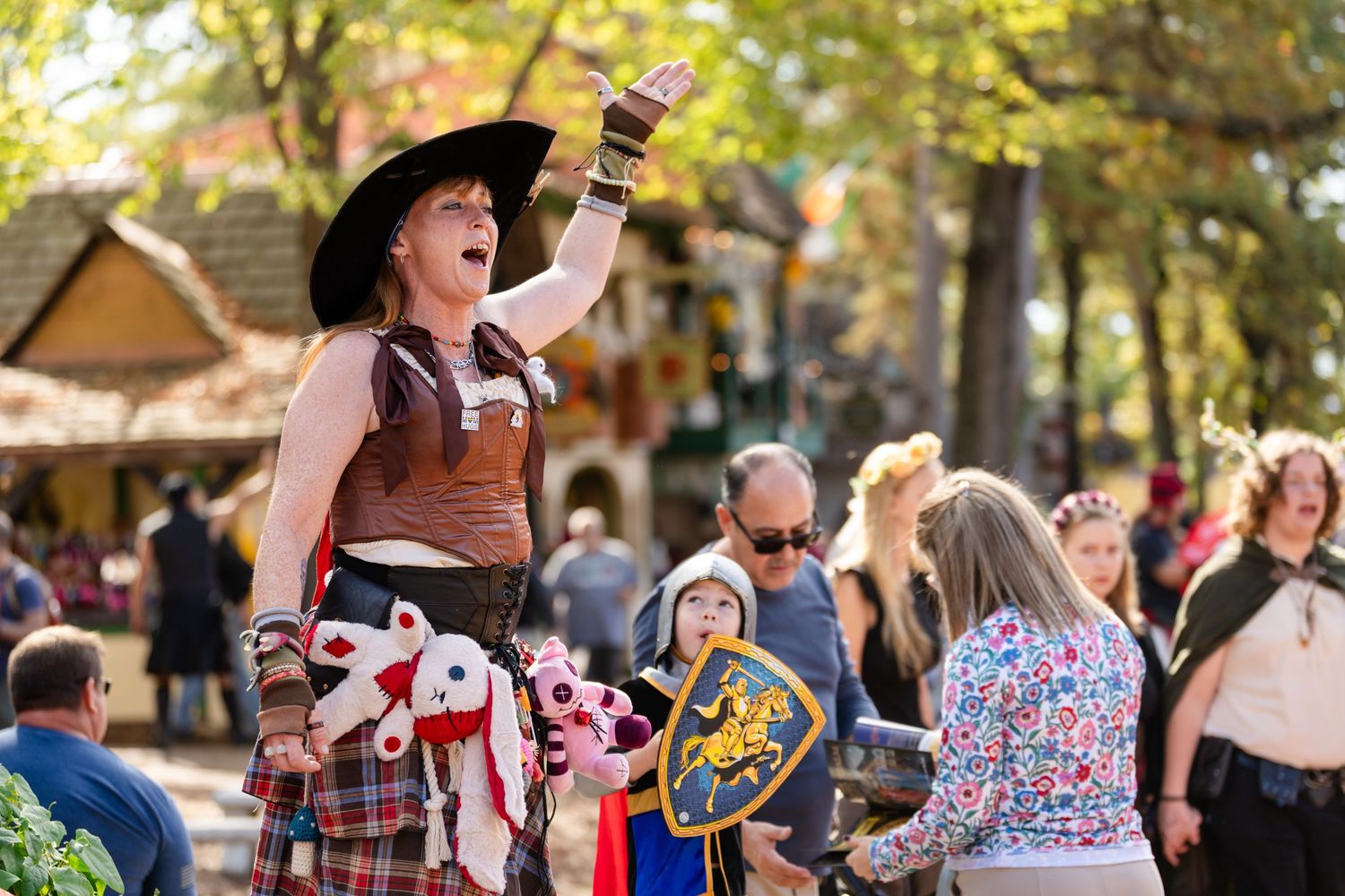 A performer in medieval costume raises a drink at a Renaissance festival with crowd in background.