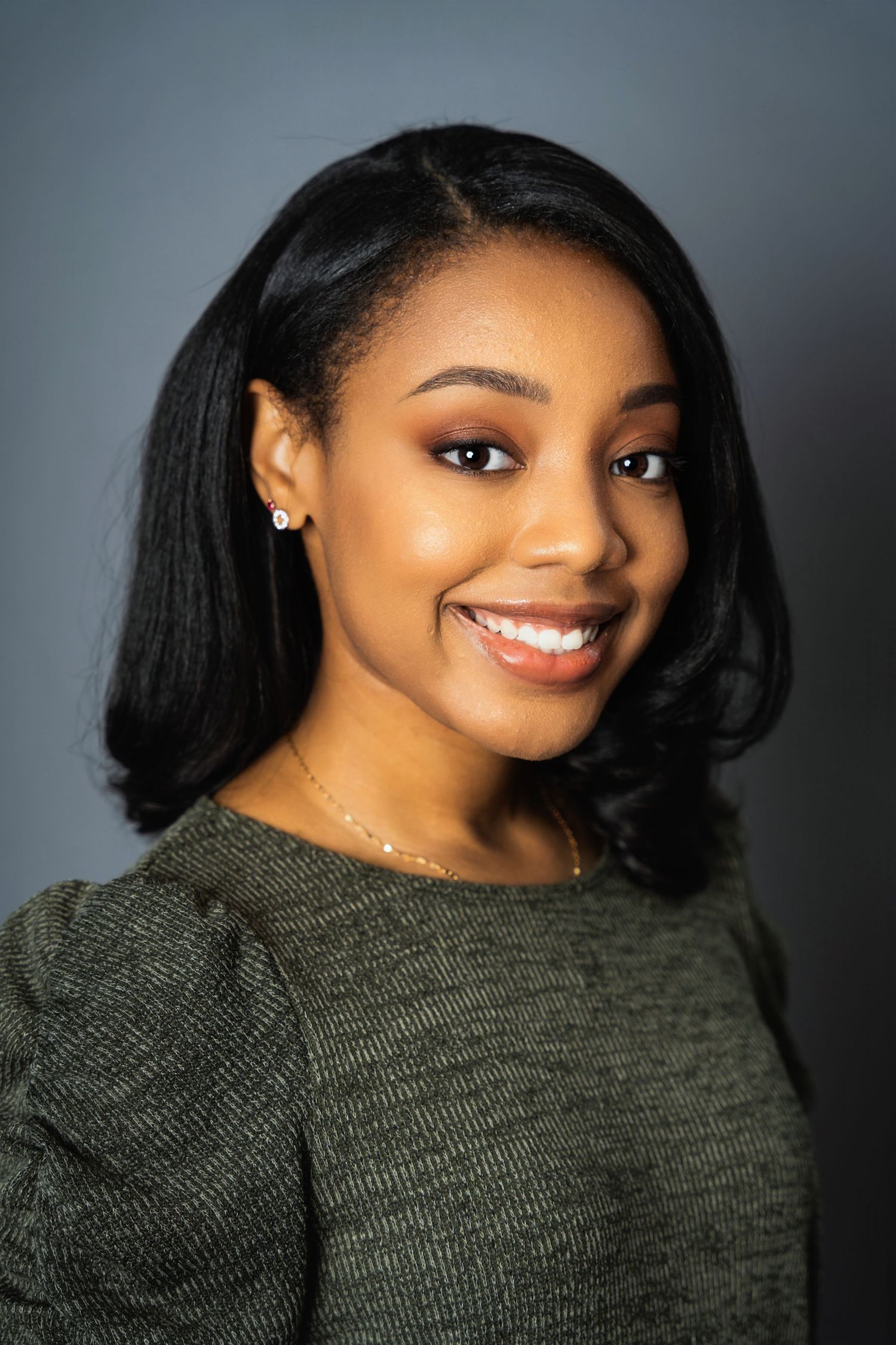 Professional studio portrait of a young woman in a grey sweater smiling warmly against a grey backdrop.