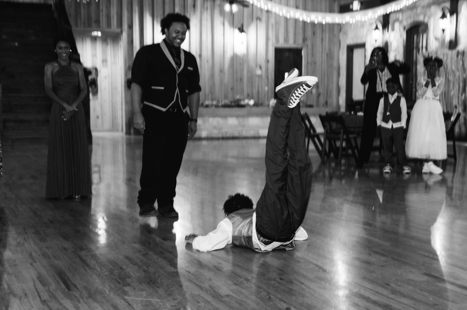 A black and white photo captures playful dancing moments at a wedding reception on a wooden floor.