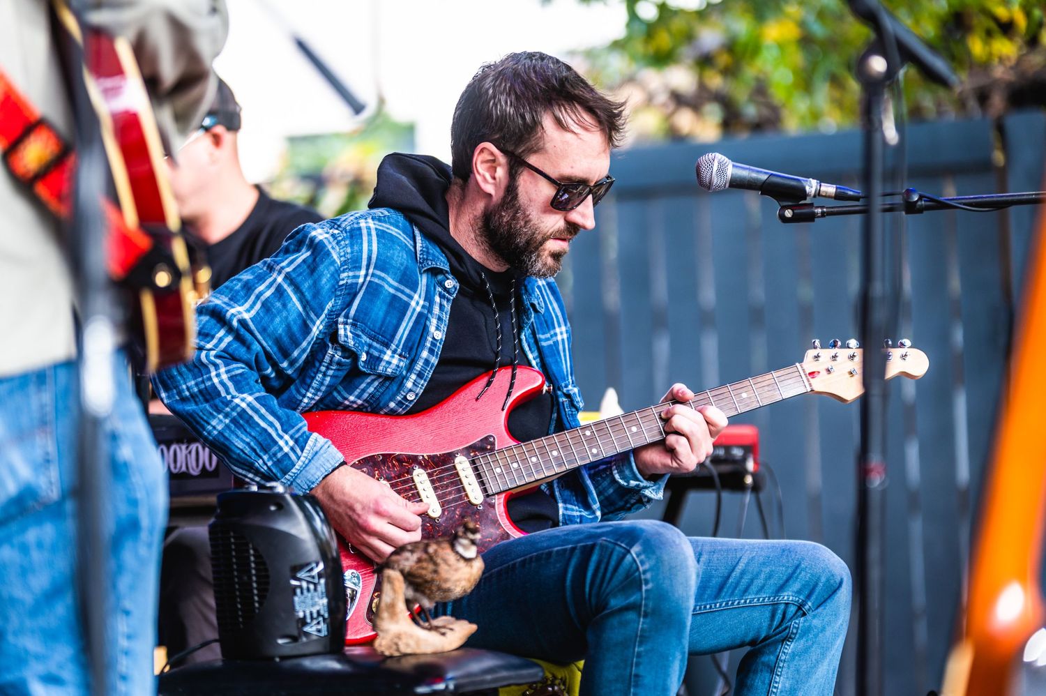 Guitarist in blue plaid shirt plays red electric guitar during outdoor performance.
