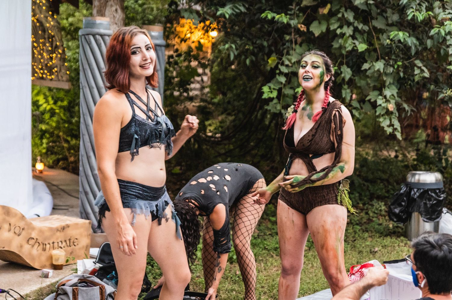 Three performers in black costumes prepare backstage in an outdoor setting with lush greenery.