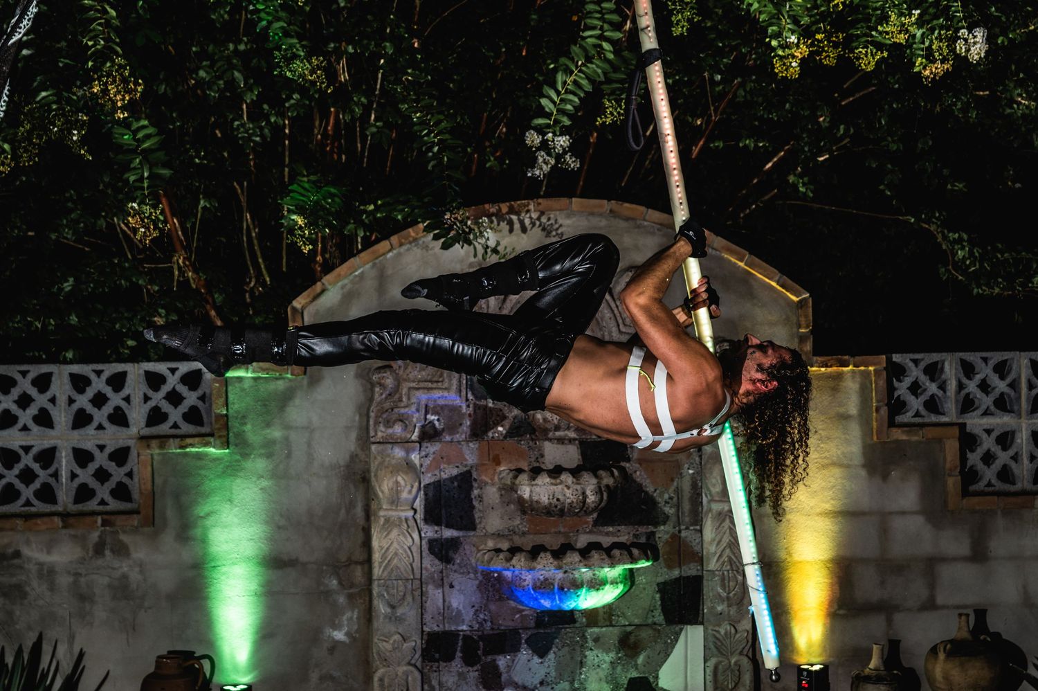 Aerial artist performs dramatic inverted pose on pole with colorful lighting effects.