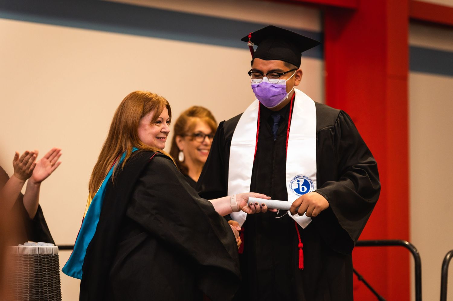 Graduate receiving diploma during graduation ceremony wearing face mask and academic regalia.