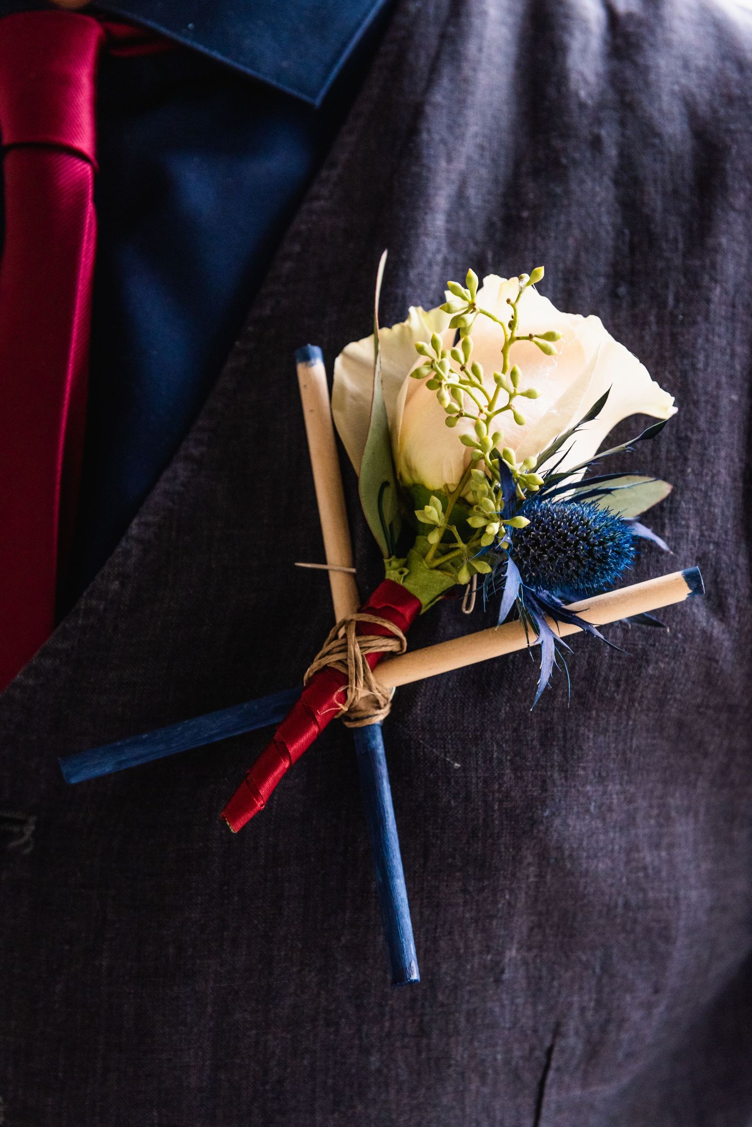 A boutonniere with a white rose, blue thistle, and greenery wrapped with red and blue ribbon on a dark jacket.