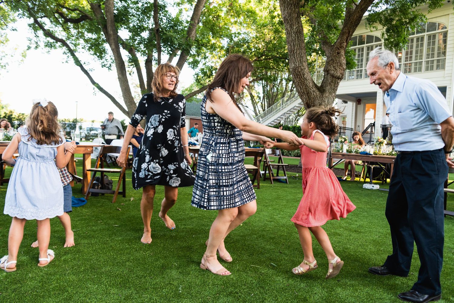 Multi-generational group dancing in a circle on grass at an outdoor celebration.
