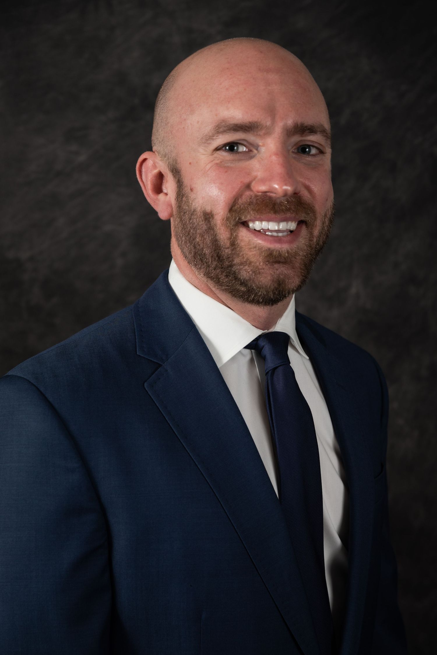 Corporate headshot with person wearing navy suit against dark backdrop.