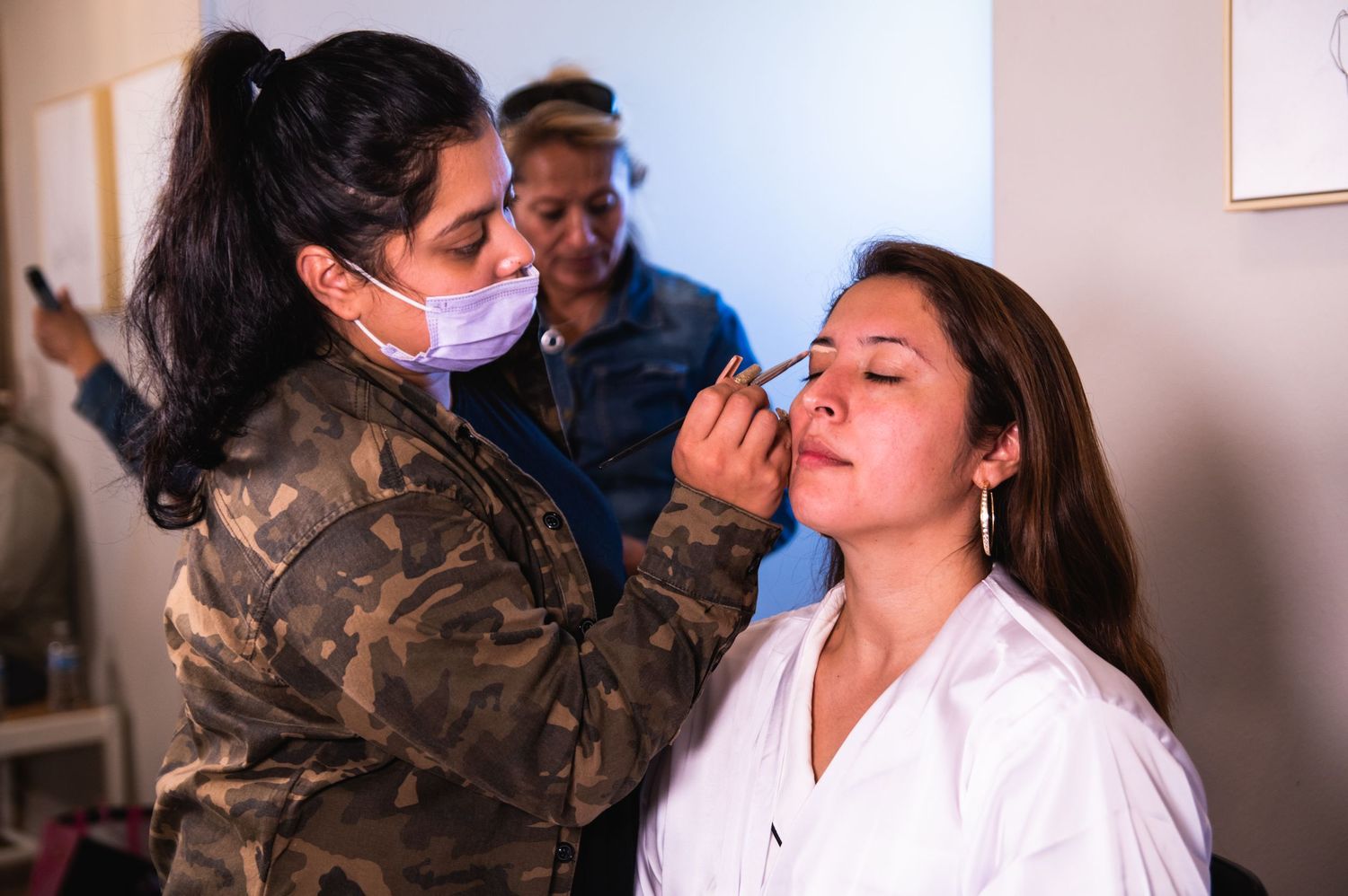 Makeup artist wearing camo jacket applies cosmetics on client wearing white robe during wedding preparation.