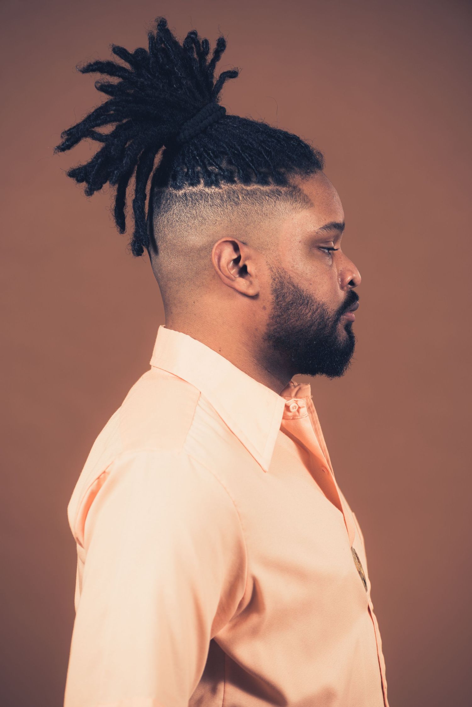Side profile portrait showing styled dreadlocks and peach colored shirt against brown backdrop.
