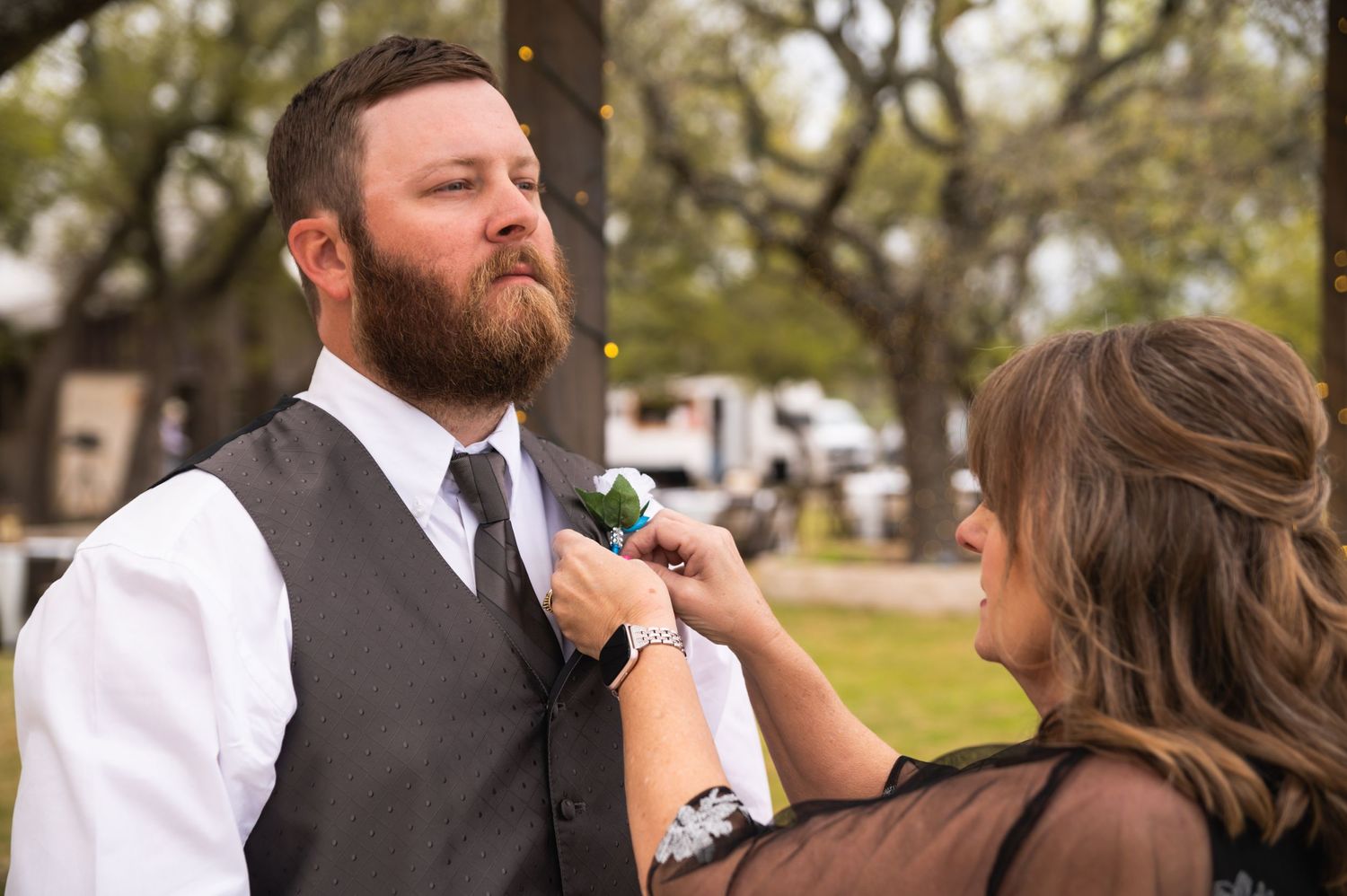 Person adjusts boutonniere on groom's grey vest during wedding preparations.
