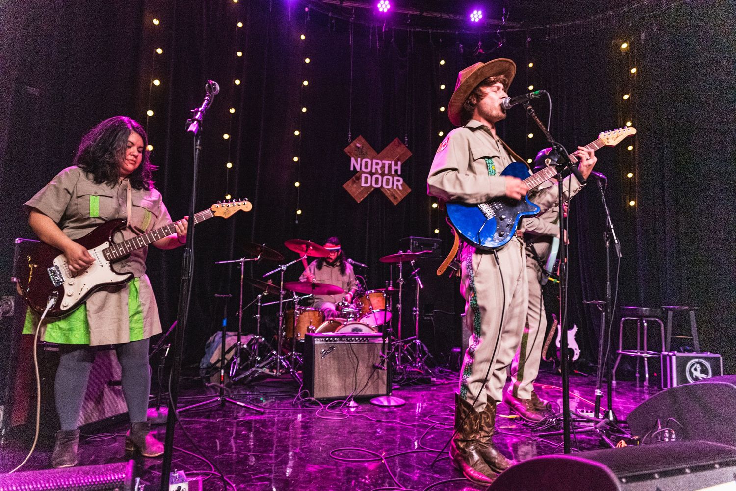 Band performs on illuminated stage with drum kit and multiple guitarists in matching outfits.