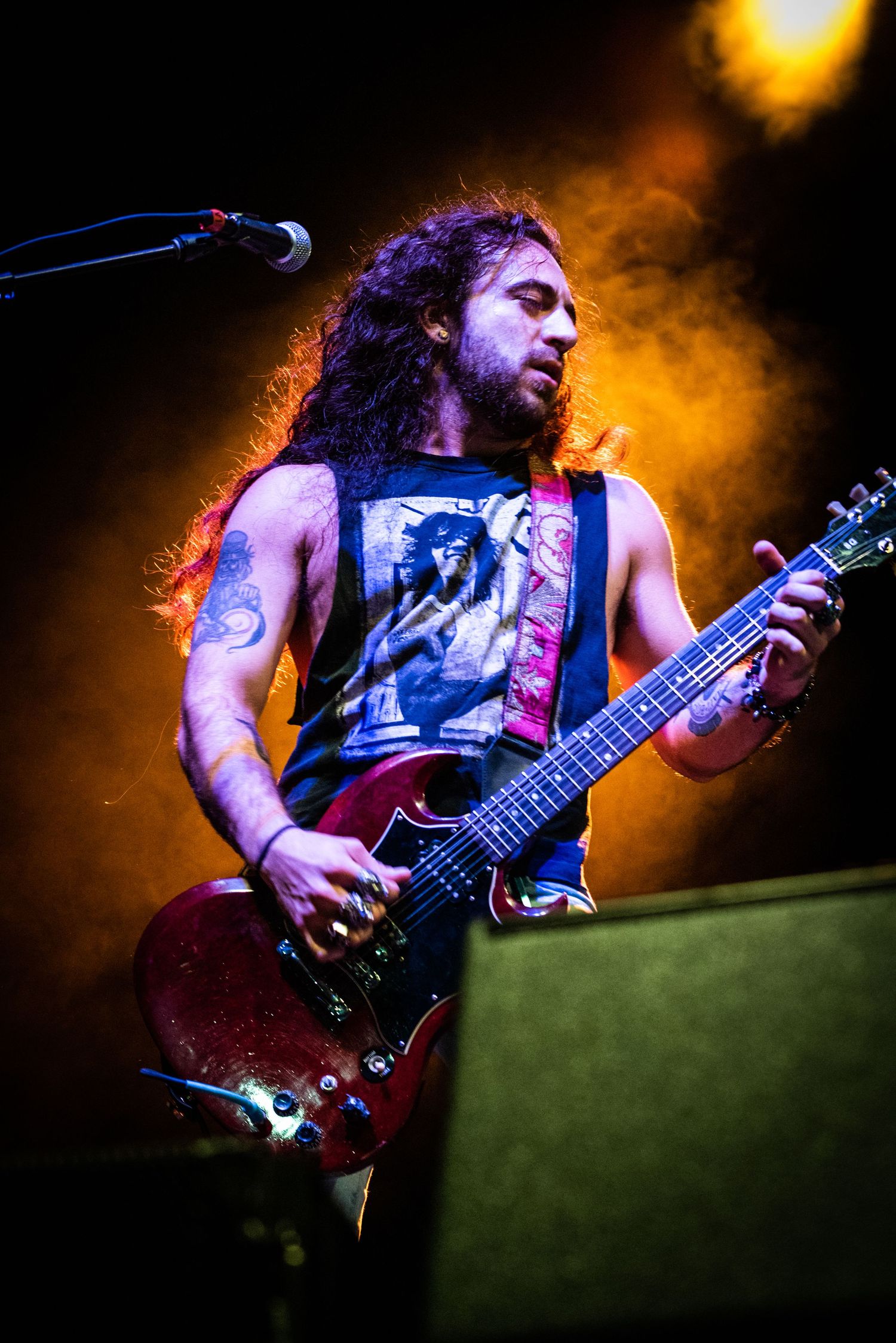 Guitarist performing with long hair and tank top against orange stage lighting.